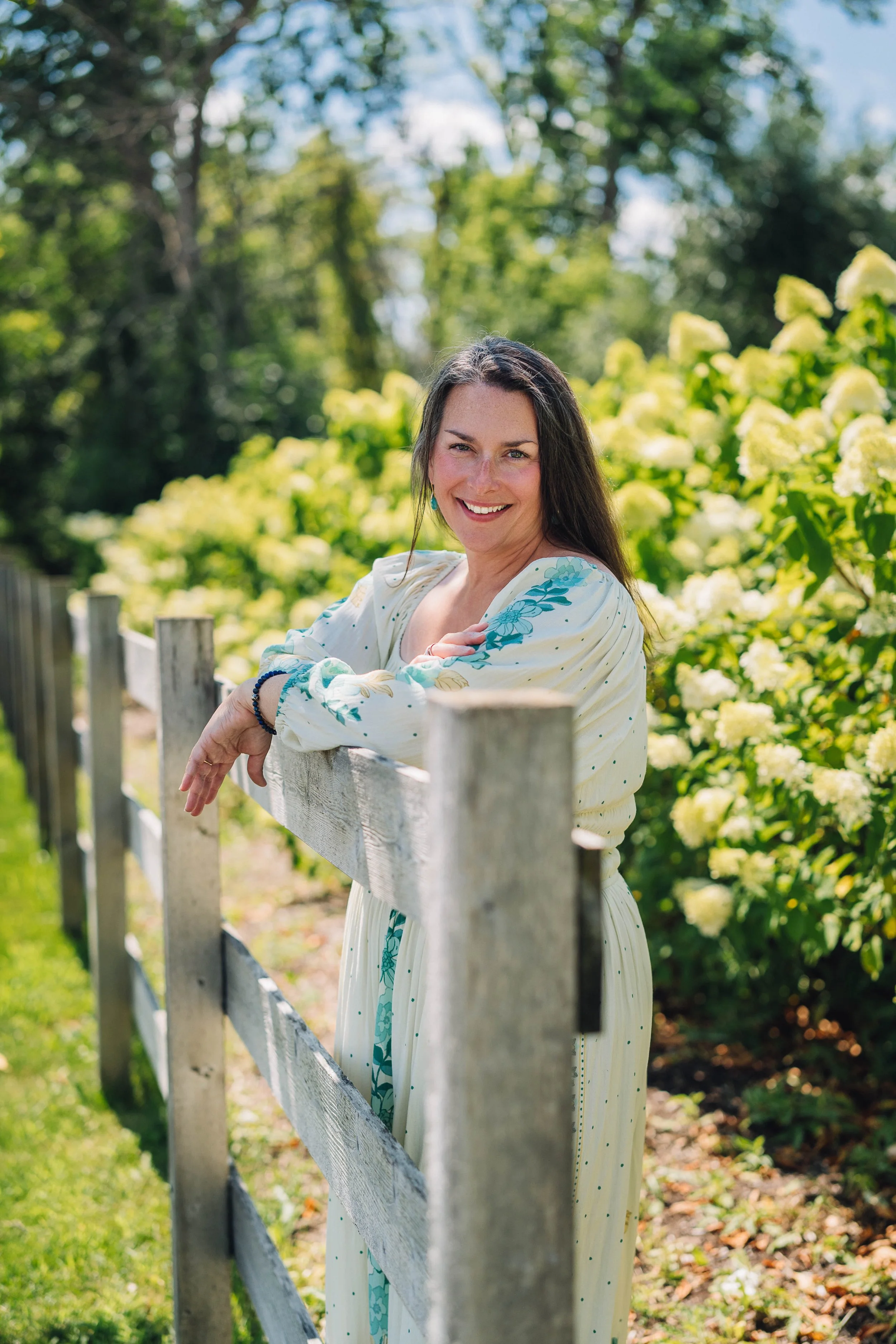 Aubree Guiffre, perimenopause and menopause coach, smiling and leaning on a wooden fence outdoors with green trees and white hydrangea flowers in the background.