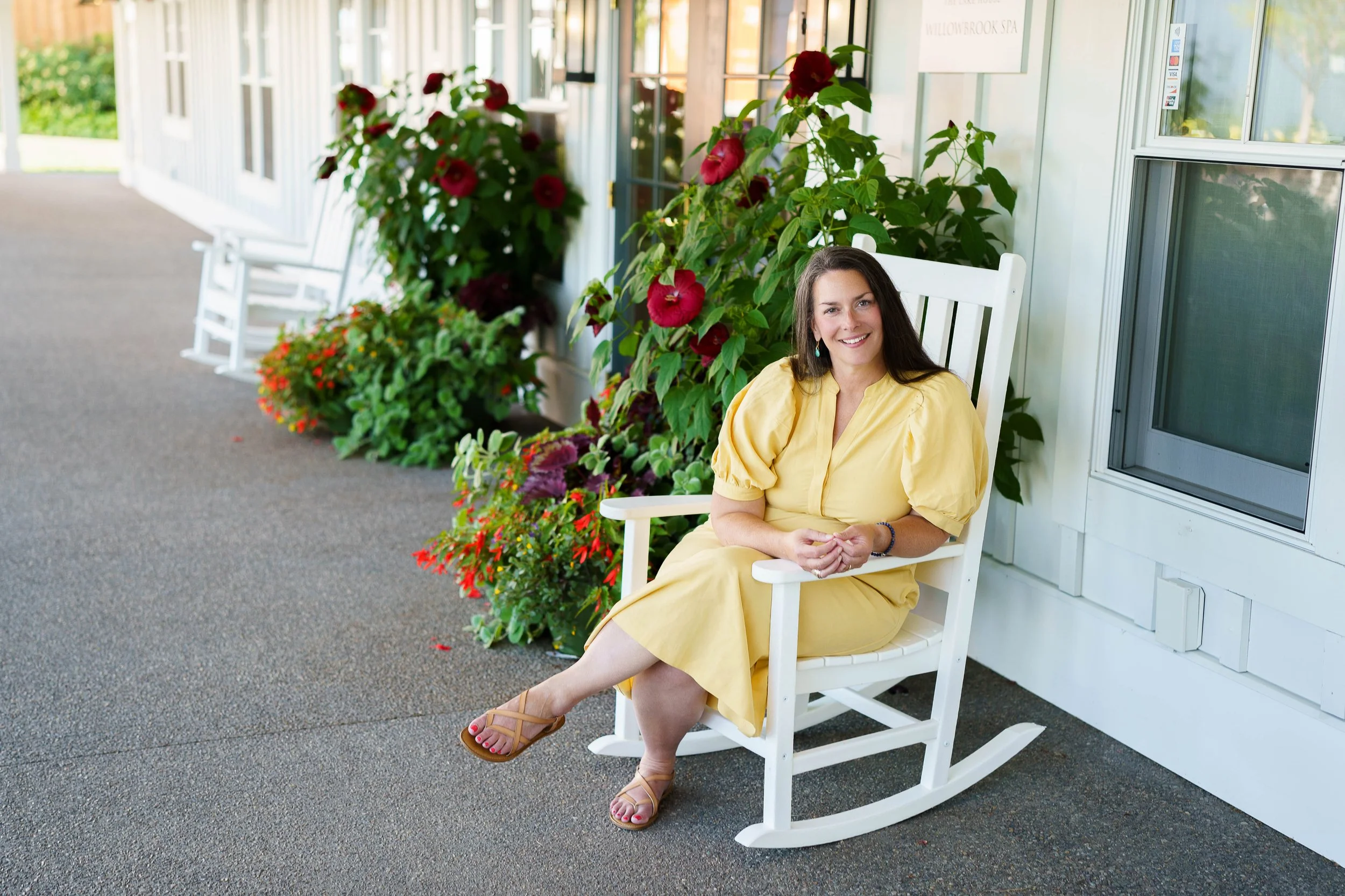 Aubree Guiffre, perimenopause and menopause coach in a yellow dress sitting on a white rocking chair outside on a porch, surrounded by colorful flowers and green plants.