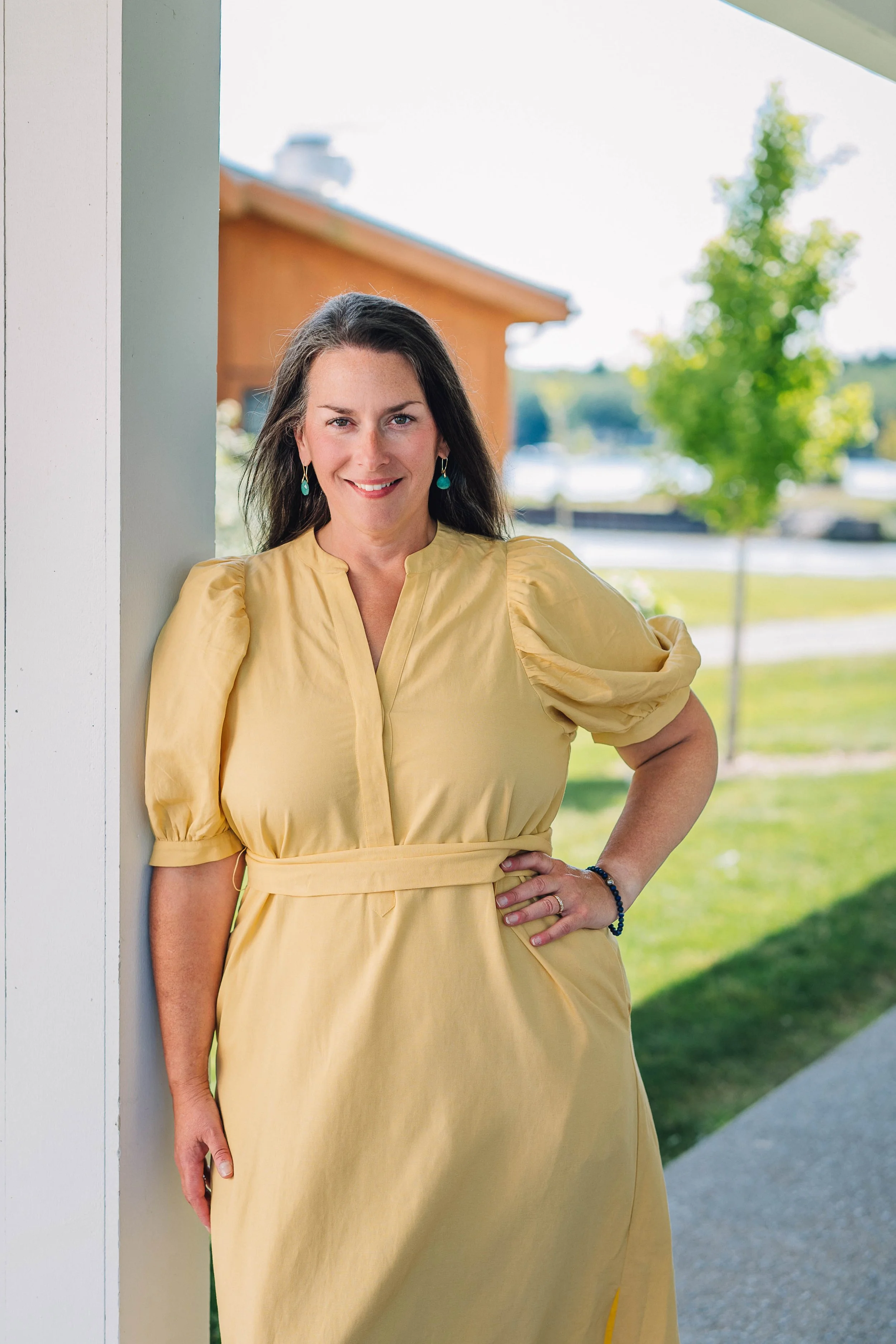 Aubree Guiffre, perimenopause and menopause coach, wearing a yellow dress with puffed sleeves and turquoise earrings, stands outdoors next to a white building, smiling at the camera.