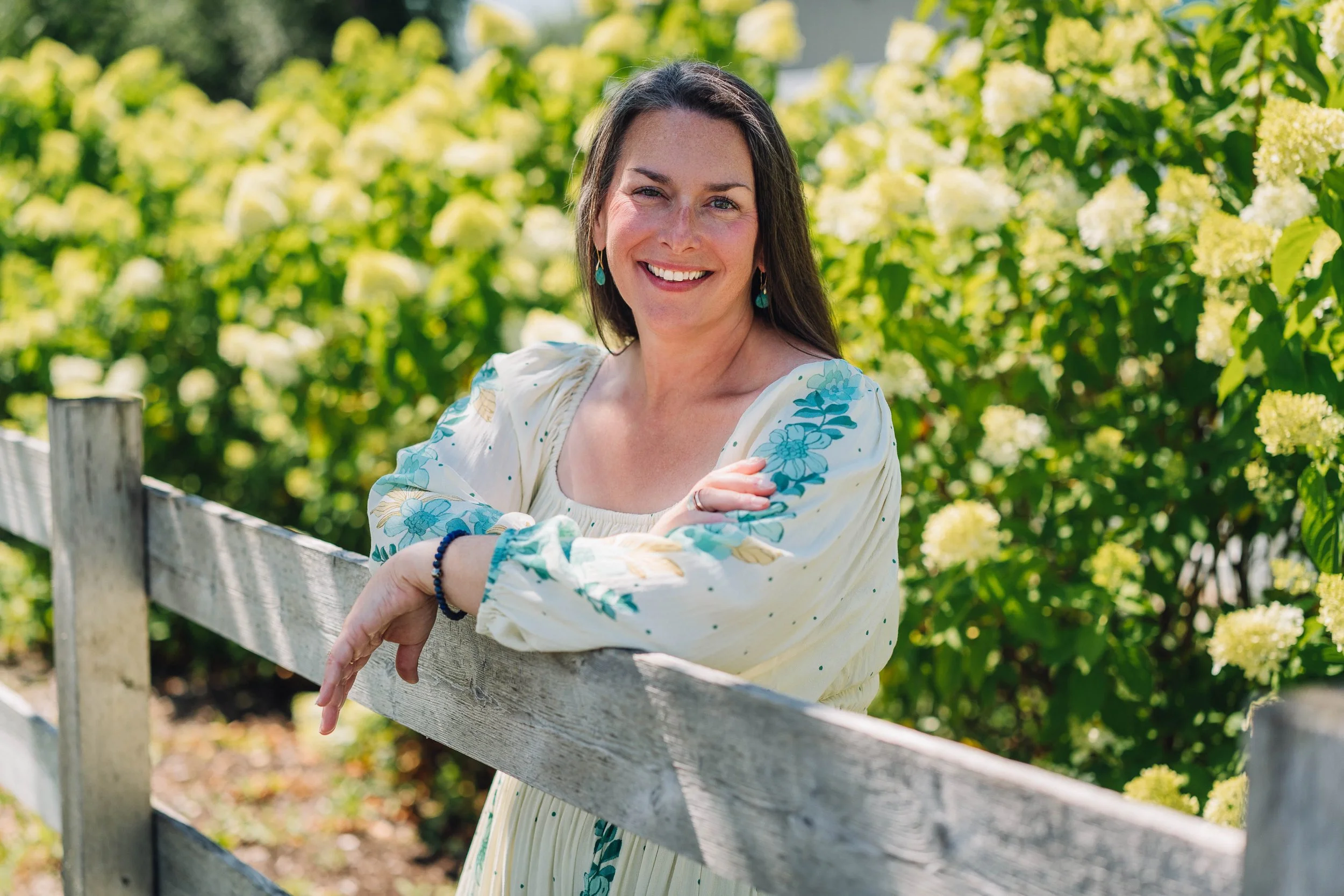 Aubree Guiffre, perimenopause and menopause coach, standing outdoors near a wooden fence, surrounded by blooming white hydrangea flowers, wearing a light-colored dress with blue floral embroidery.