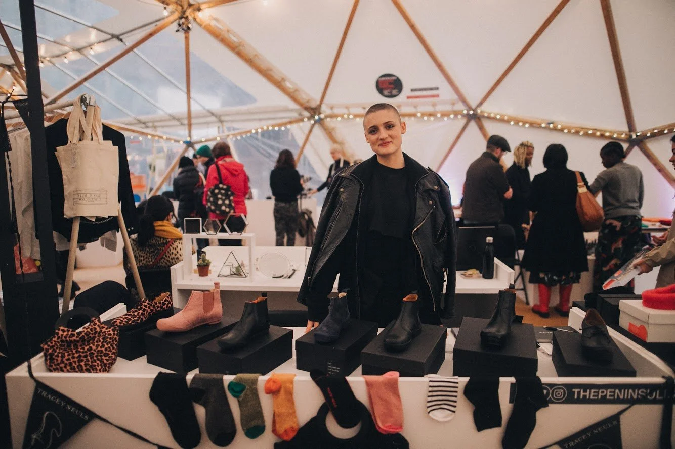 A young woman with a shaved head wearing a black leather jacket standing behind a display of shoes and socks at a market or fair inside a tent with wooden support beams and string lights.