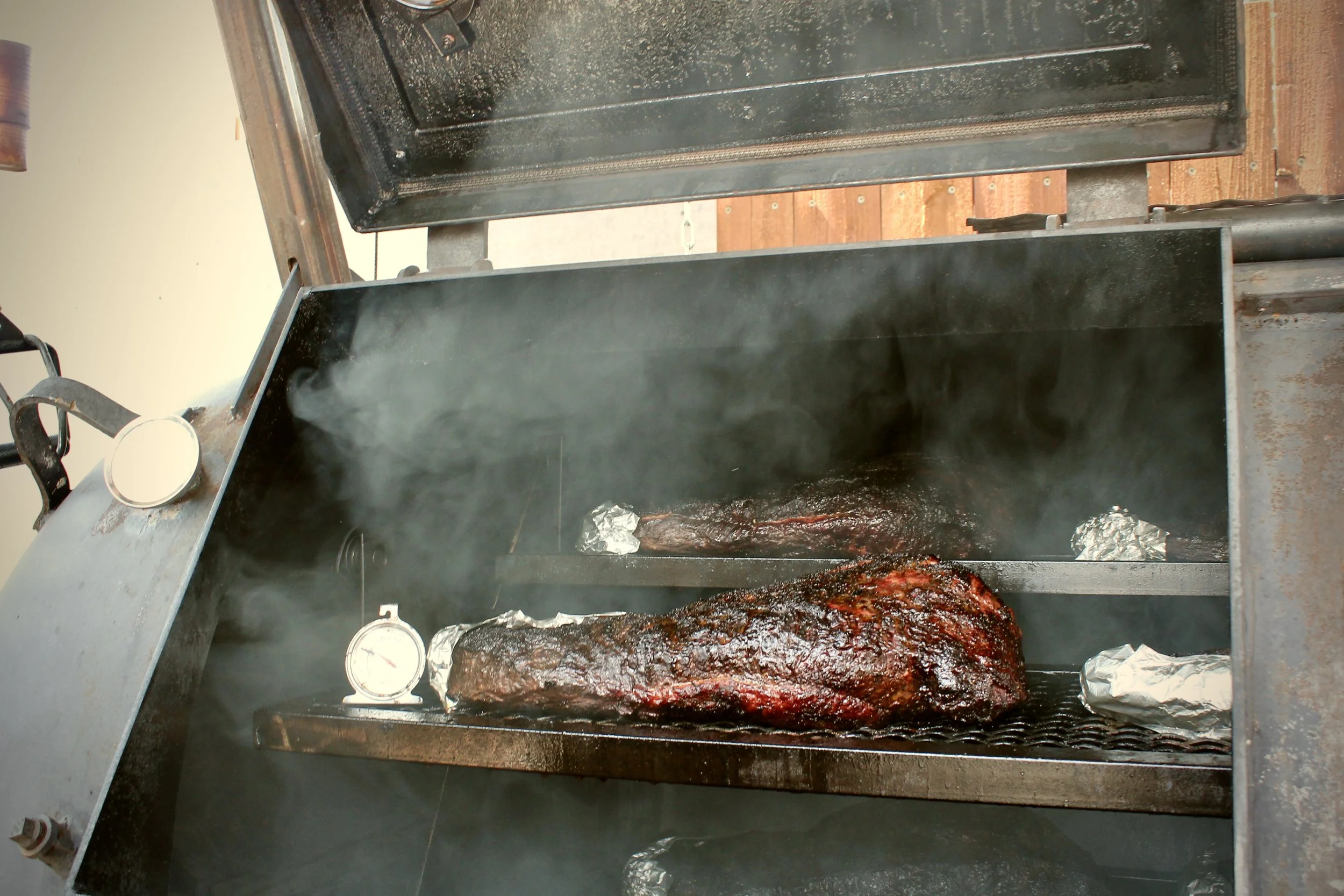 A large piece of smoked meat cooking inside a smoker, with smoke billowing out and temperature gauges attached.
