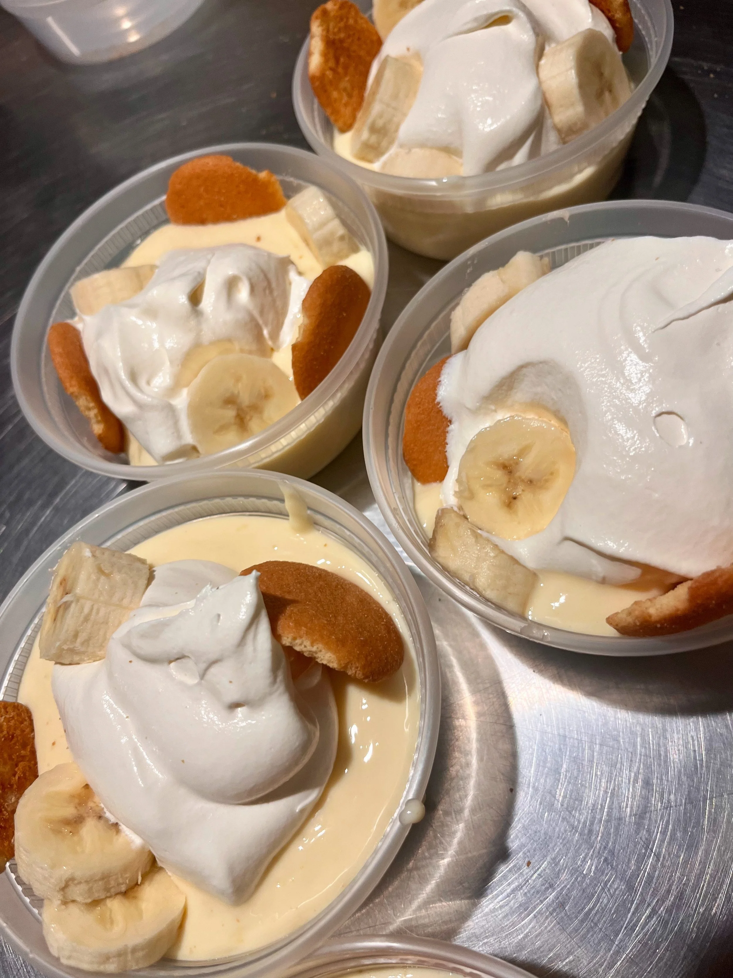 Four small cups of banana pudding topped with whipped cream, banana slices, vanilla wafers, and cookie pieces on a metal surface.