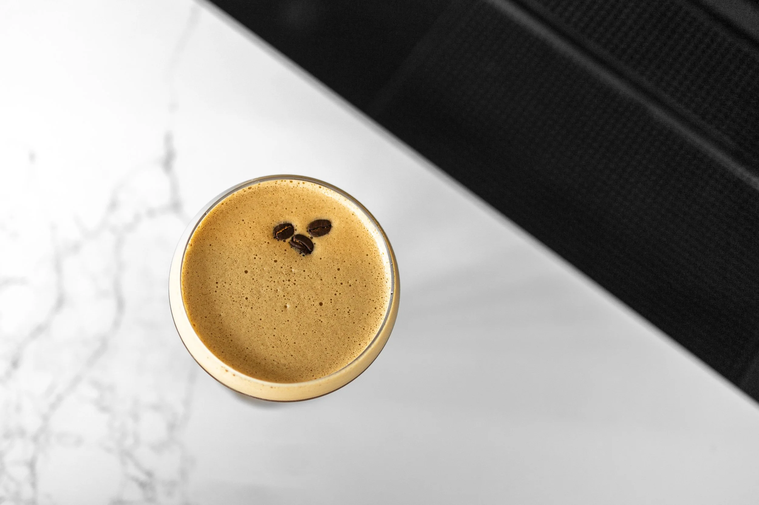 Top view of a glass of iced coffee with coffee beans floating on top, placed on a white surface with a textured black object in the background.