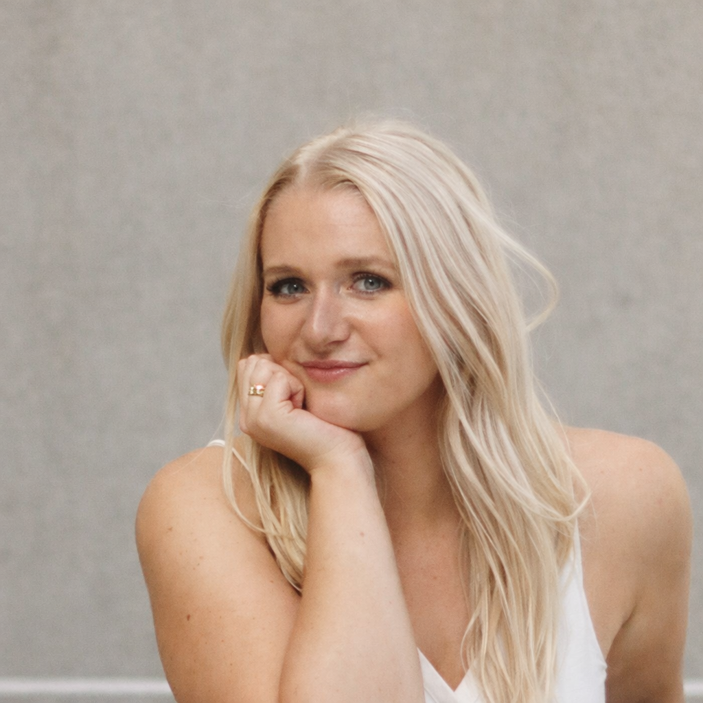A woman with long blonde hair and blue eyes, resting her chin on her hand, smiling gently at the camera against a neutral background.