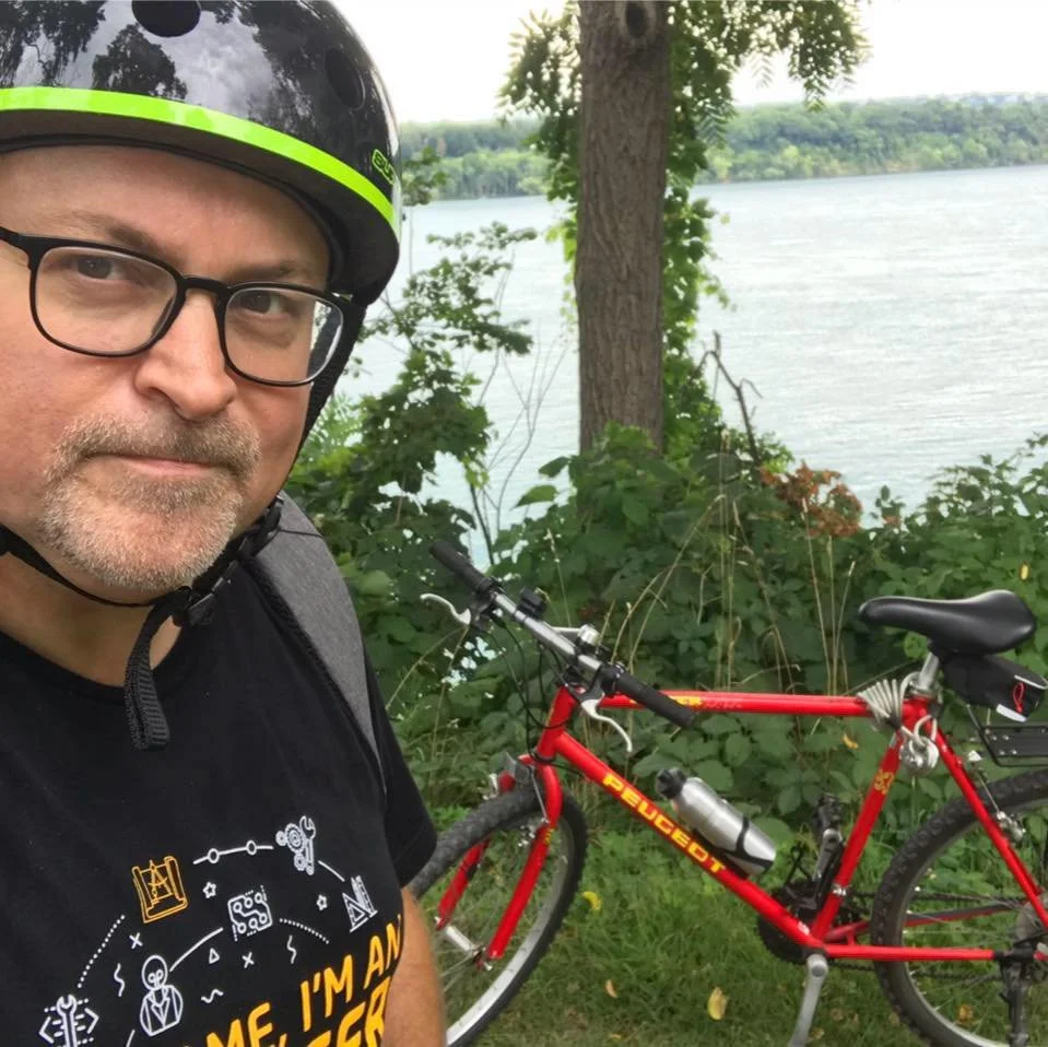 A man with glasses and a beard wearing a helmet and black t-shirt takes a selfie beside a red bicycle near a lake, with trees and hills in the background.