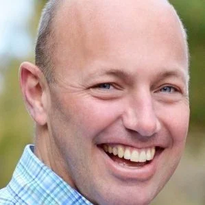 Close-up of a smiling man outdoors with a blurred green background.