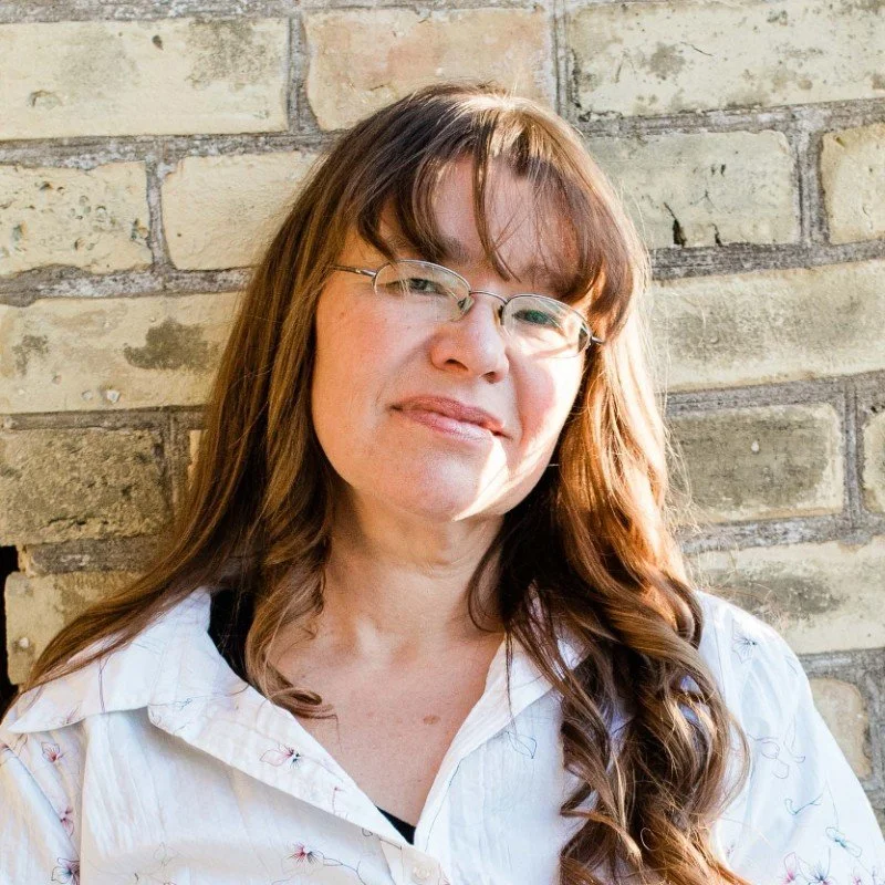 A woman with long brown hair and glasses standing against a brick wall, smiling slightly.