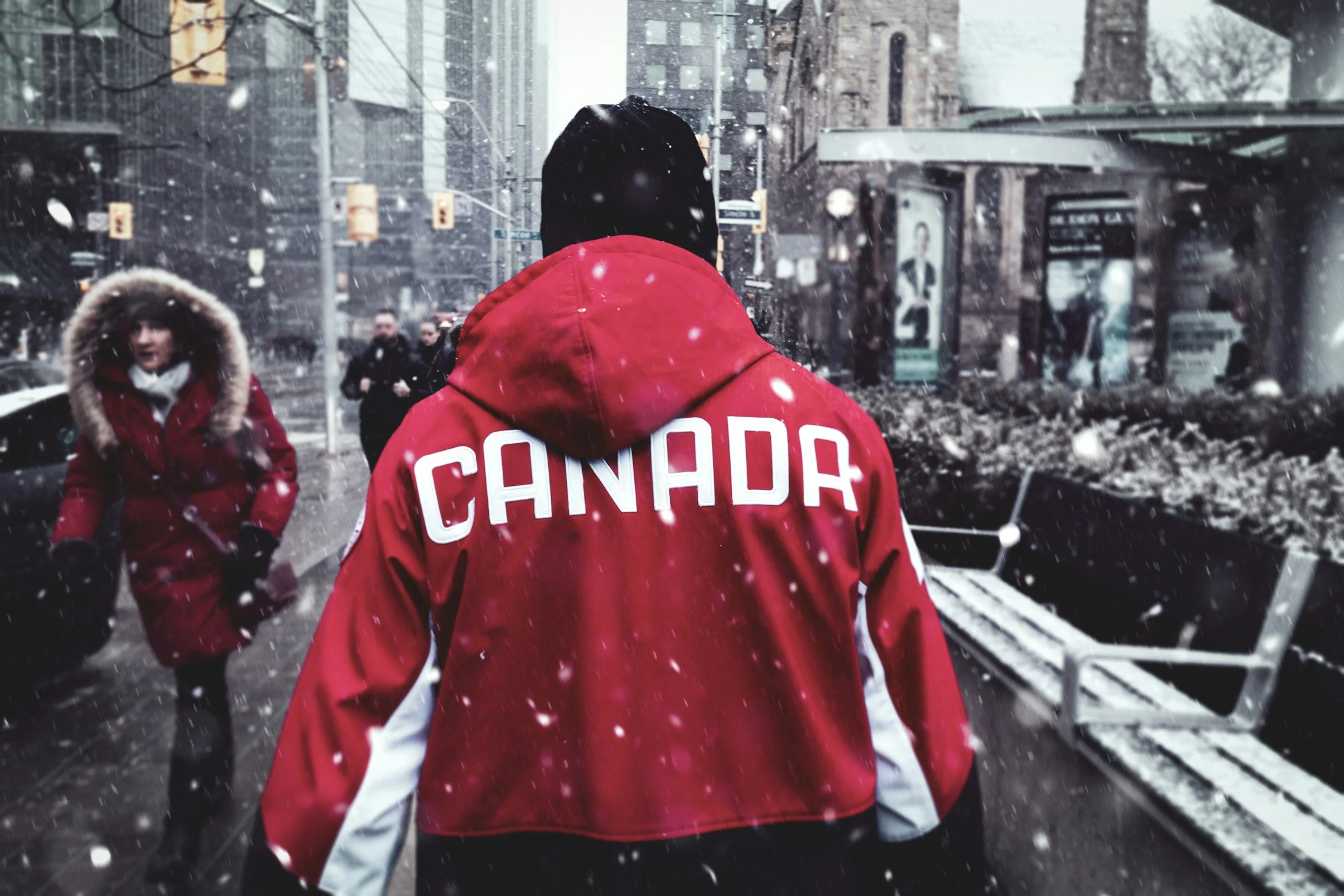A person wearing a red jacket with 'CANADA' written on the back walks in a snowy city street, with a snowstorm in progress, surrounded by pedestrians and city buildings.