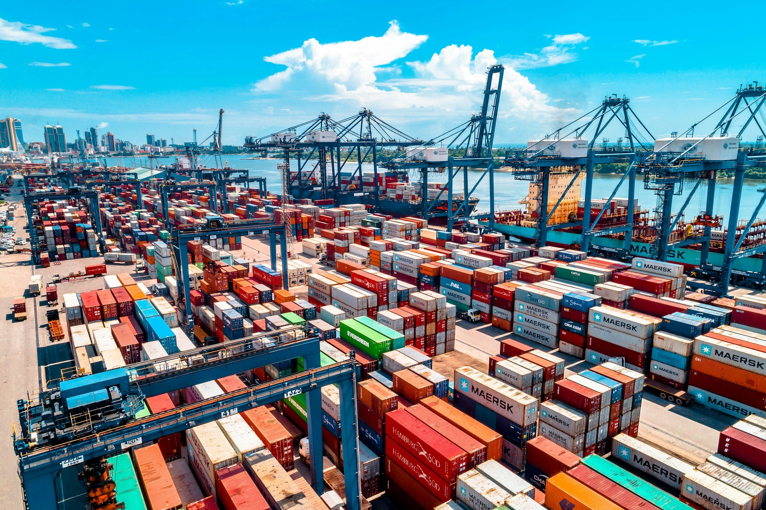 A busy container port with numerous stacked shipping containers, large cranes, and cargo ships on a river under a blue sky with some clouds.