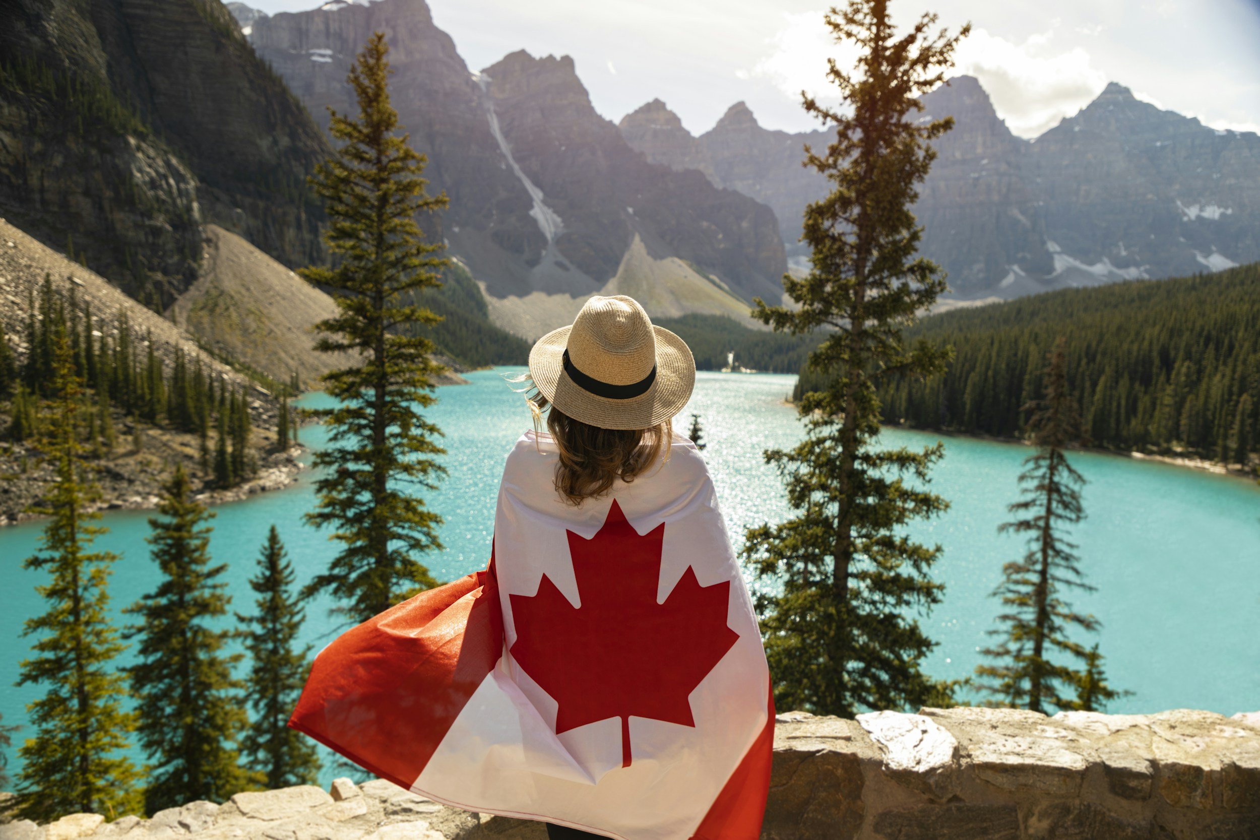 A woman with a hat and a Canadian flag draped over her shoulders is sitting on a stone wall, overlooking a turquoise lake surrounded by pine trees and mountains.