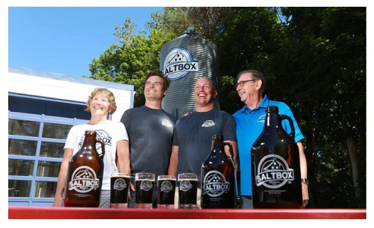 Four people standing behind a table with bottles and glasses of beer, outdoors with trees and a large salt container in the background.