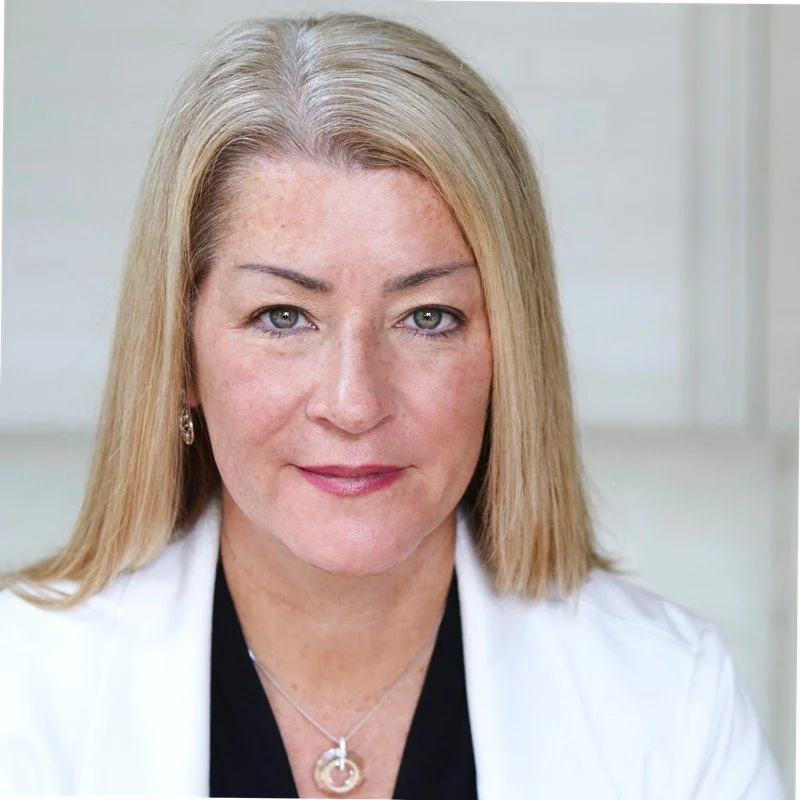 A woman with blonde hair, fair skin, and light-colored eyes, wearing a white coat and a necklace, posing in a professional setting.