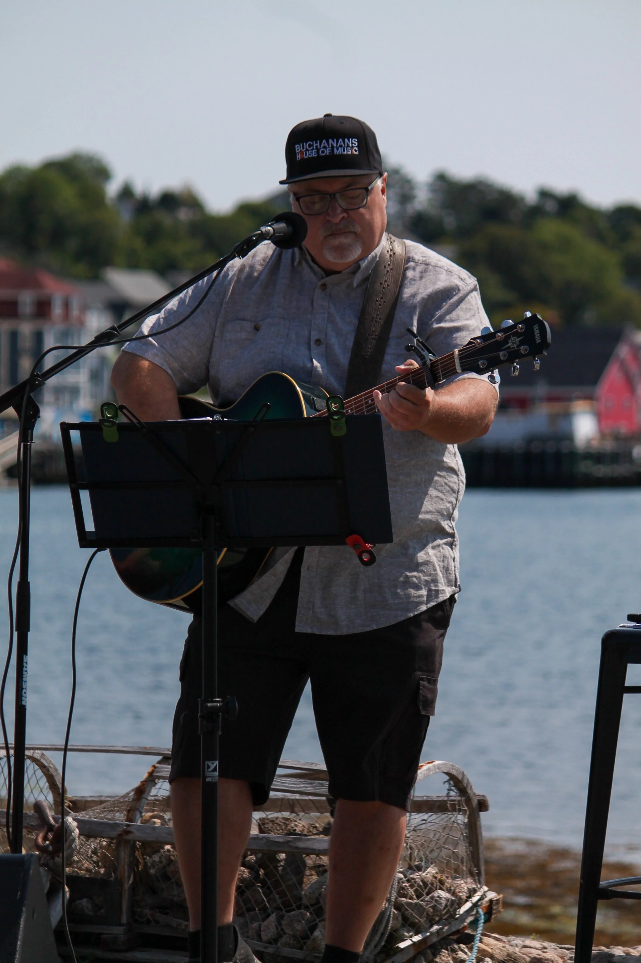 A man with glasses and a cap playing an acoustic guitar on a waterfront stage with houses in the background.