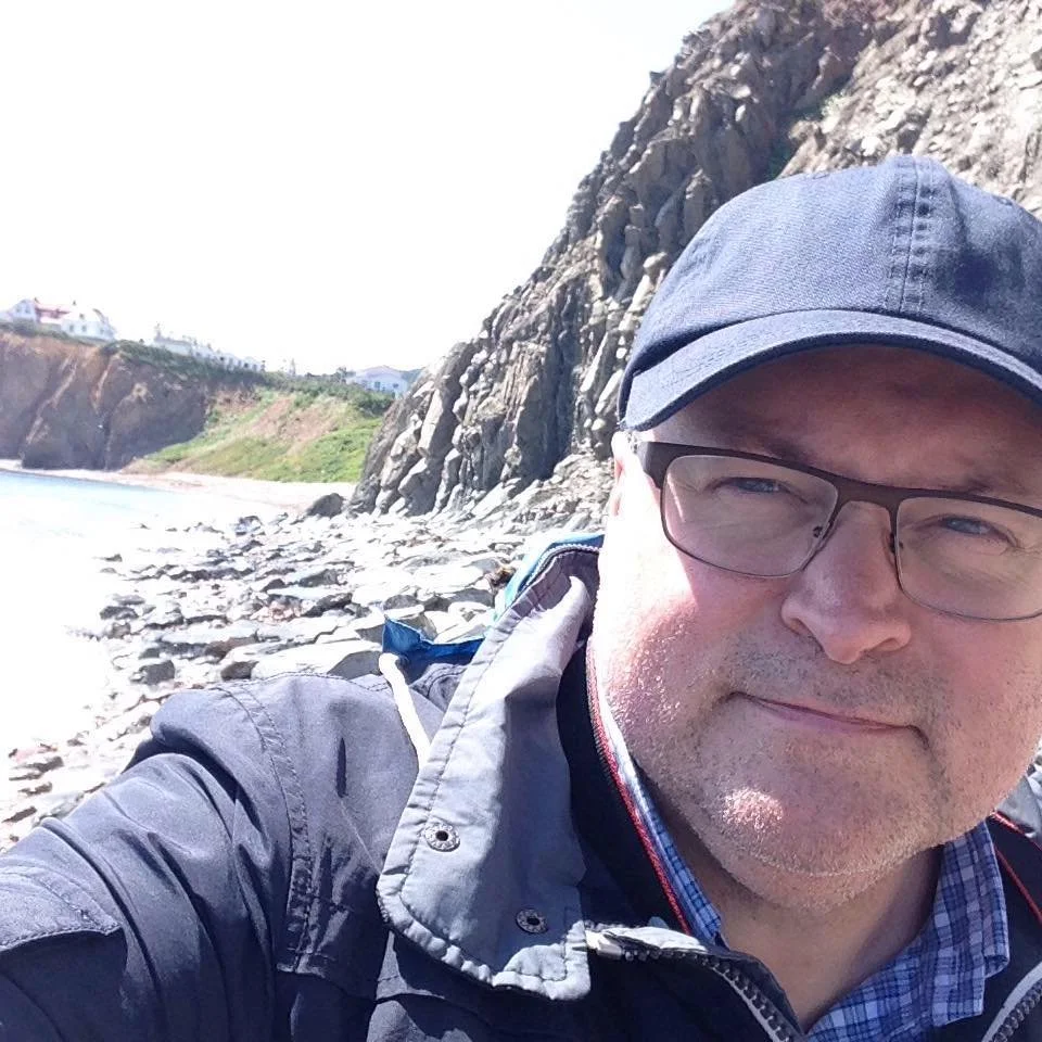 A man wearing glasses and a cap takes a selfie outdoors near a rocky shoreline with cliffs and houses in the background during daytime.