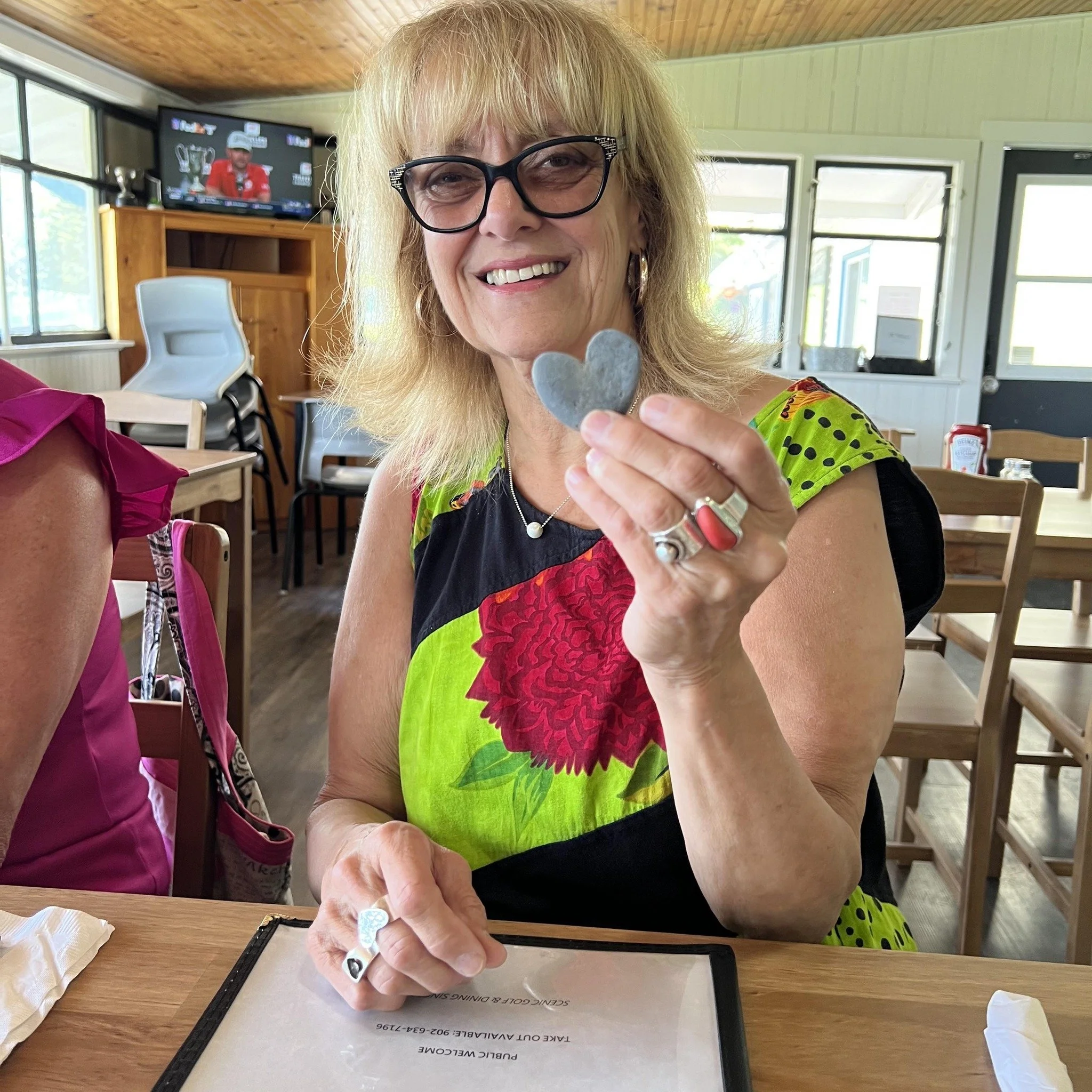 A woman with blonde hair and glasses, smiling, holding two small heart-shaped stones in front of her, sitting in a restaurant.