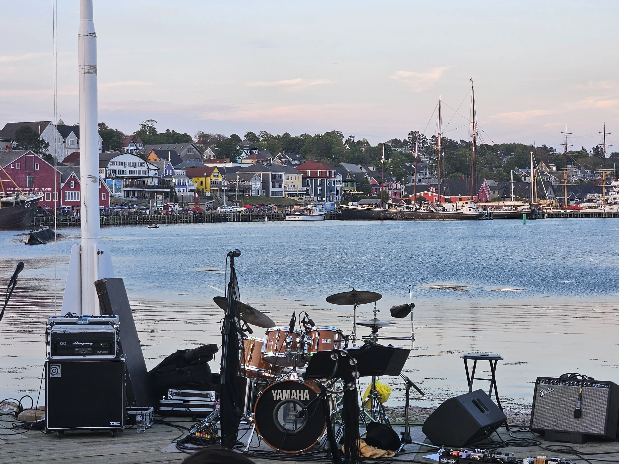 Musical instruments and speakers set up on a wooden dock near water, with a colorful harbor town of boats and houses in the background.