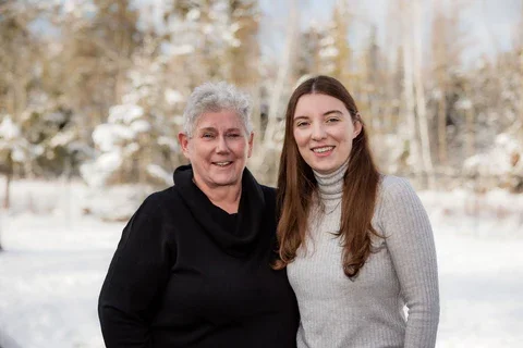 A young woman and an older woman standing outdoors in a snowy landscape, smiling at the camera.