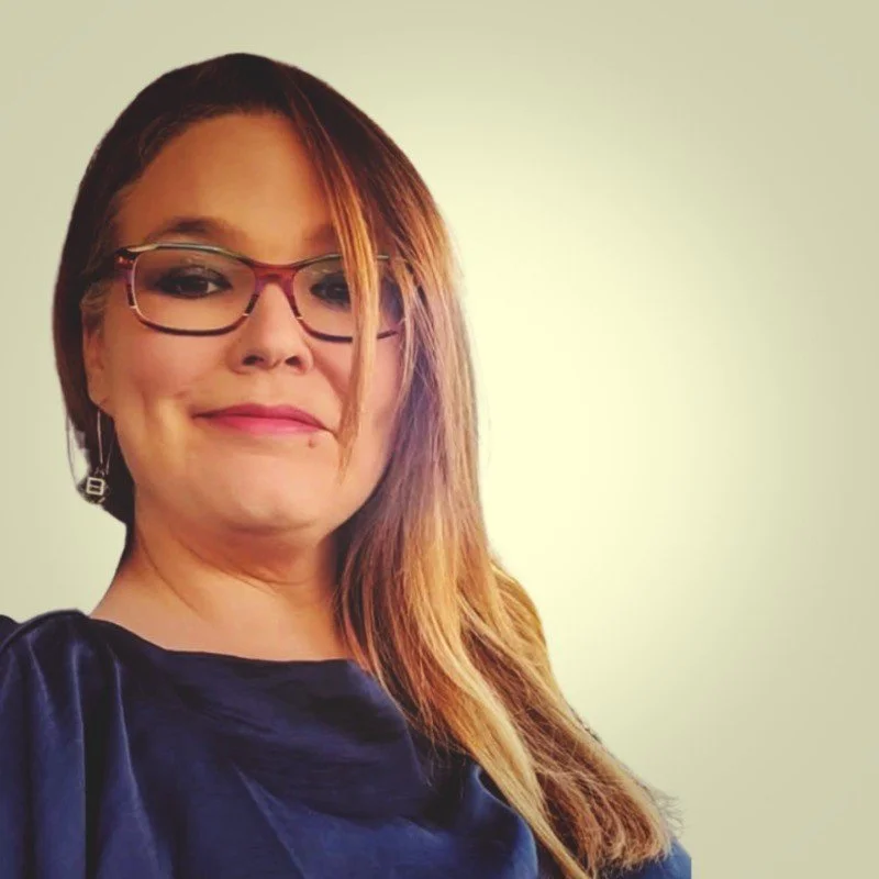 A woman with long reddish-brown hair, glasses, and earrings, smiling at the camera against a plain background.