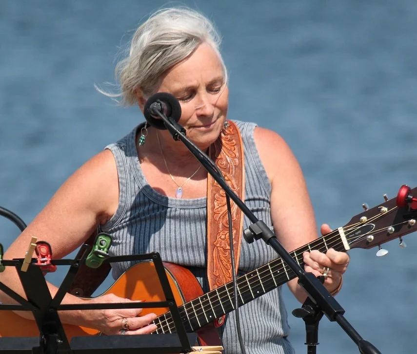An older woman with short gray hair playing an acoustic guitar and singing into a microphone outdoors near a body of water.