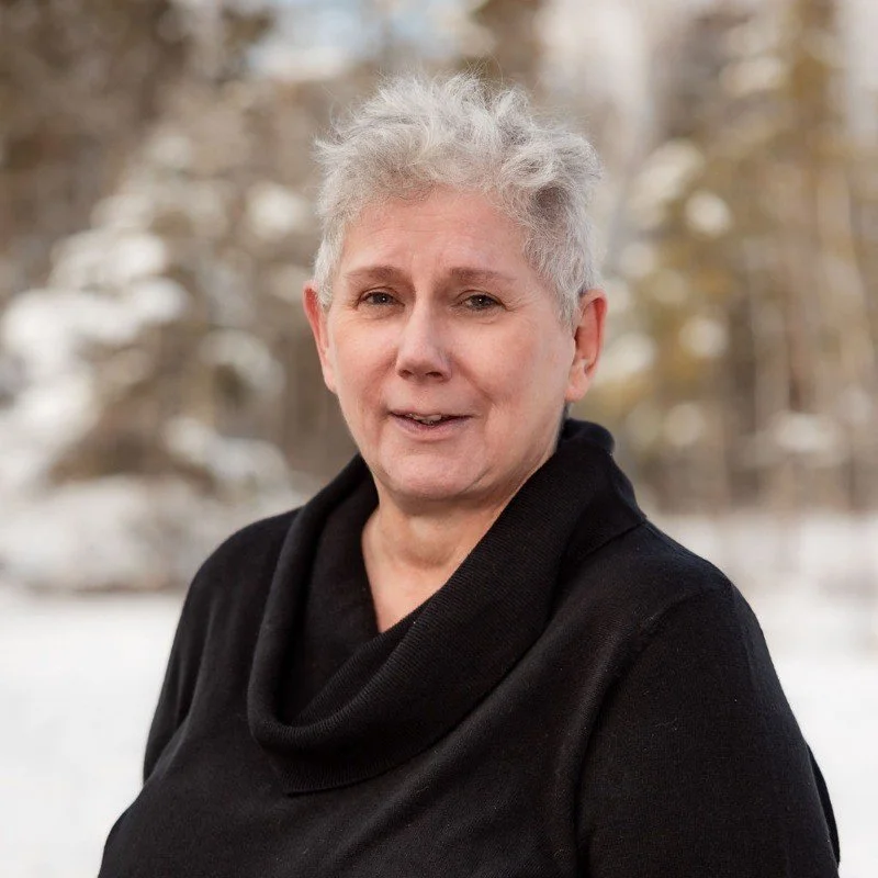 A woman with short gray hair wearing a black top standing outdoors in a winter landscape with snow-covered trees in the background.