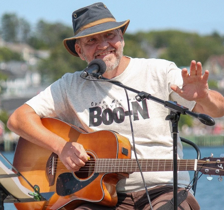 Man wearing a wide-brimmed hat, playing an acoustic guitar outdoors with water in the background, smiling and gesturing with his right hand.