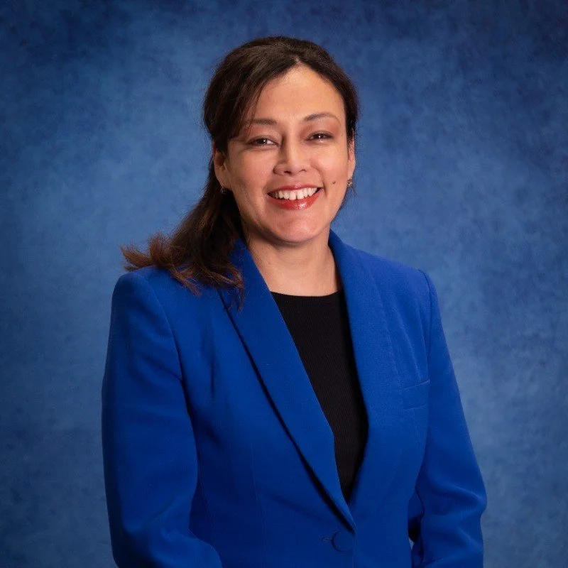 A woman with dark brown hair, smiling, wearing a bright blue blazer and black top, standing against a textured blue background.