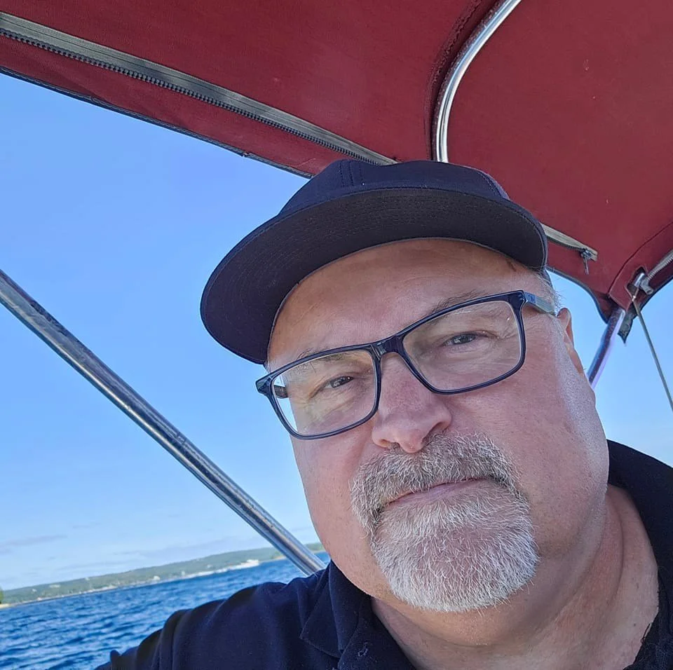A man with glasses and a cap aboard a boat, with a clear blue sky and water in the background.