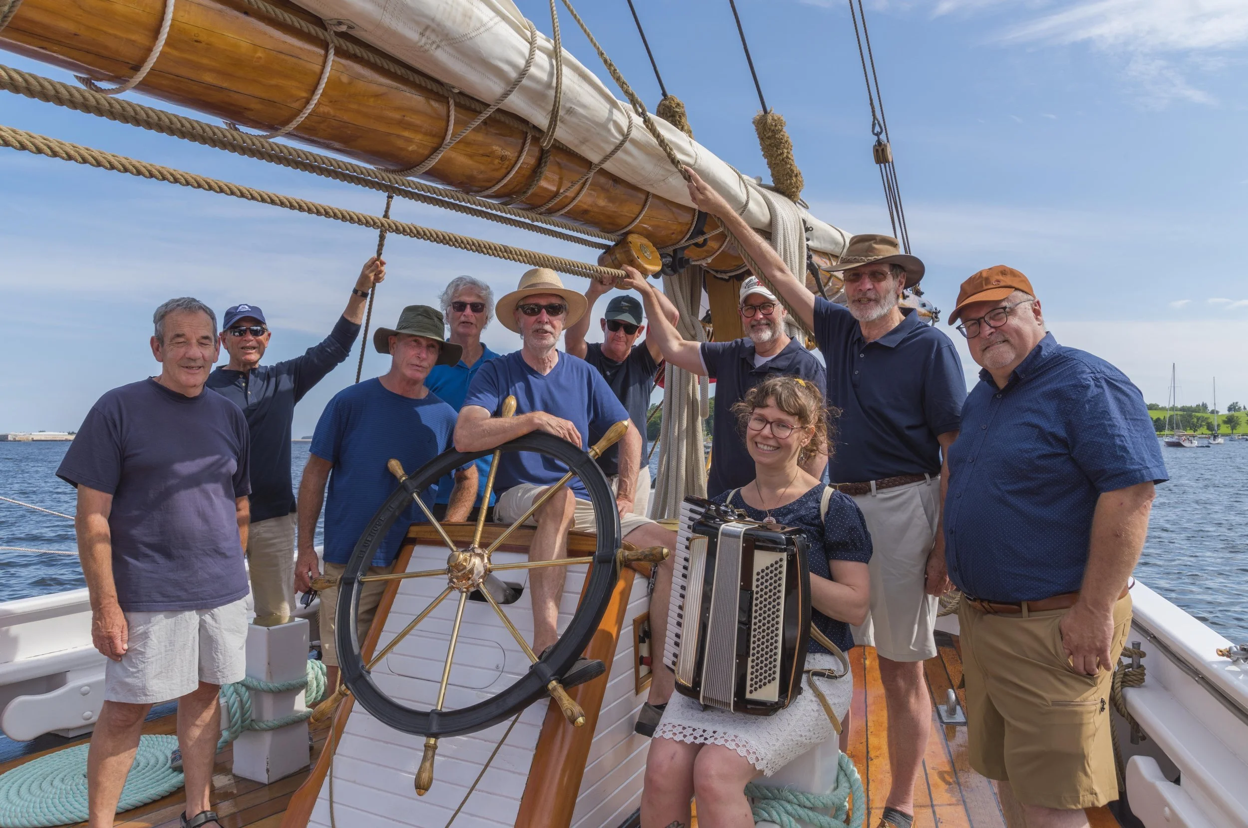 A group of nine people on a sailboat, posing for a photo on a sunny day with blue skies and water in the background. Some are wearing hats and sunglasses, and one woman is holding a small accordion. They are gathered around the ship's wheel and mast.