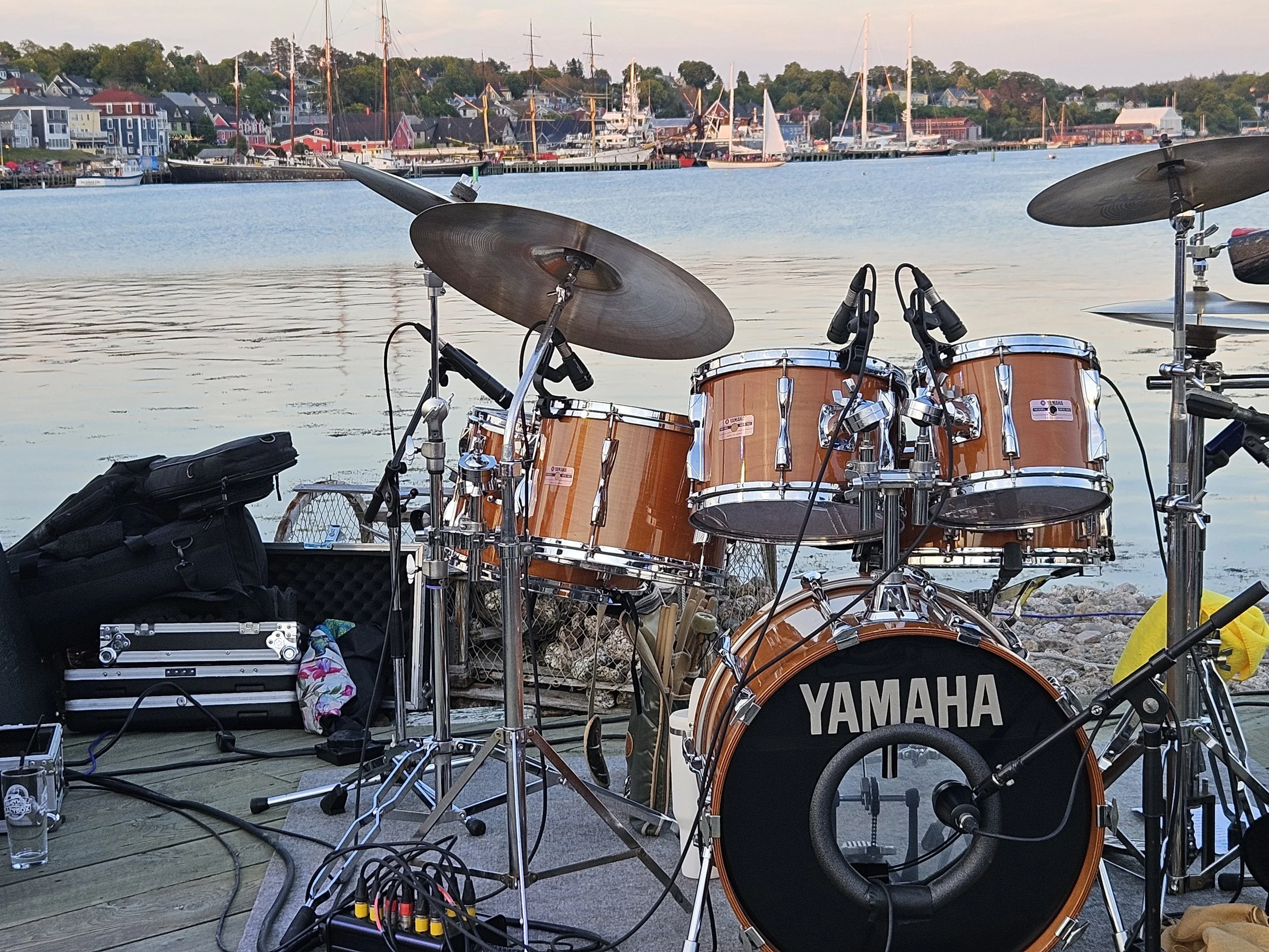 A drum set with a Yamaha bass drum, toms, cymbals, and microphones set up on a dock near a body of water with boats, houses, and a town on the opposite shore.