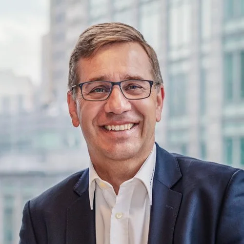 Professional man with glasses, smiling, wearing a dark suit and white shirt, standing in an office with glass windows.