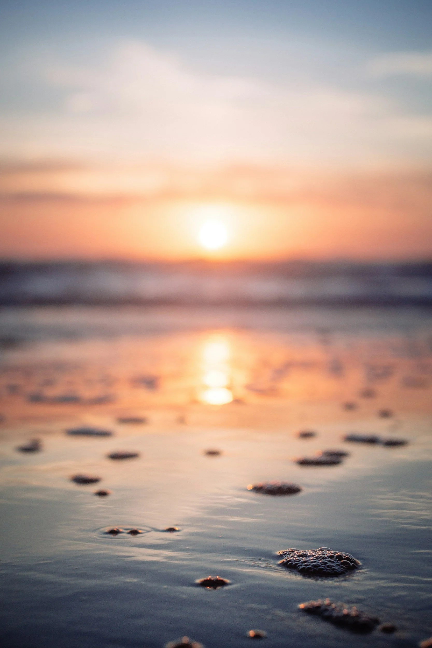 Blurred photo of a sunset over the ocean with foam bubbles on wet sand in the foreground.