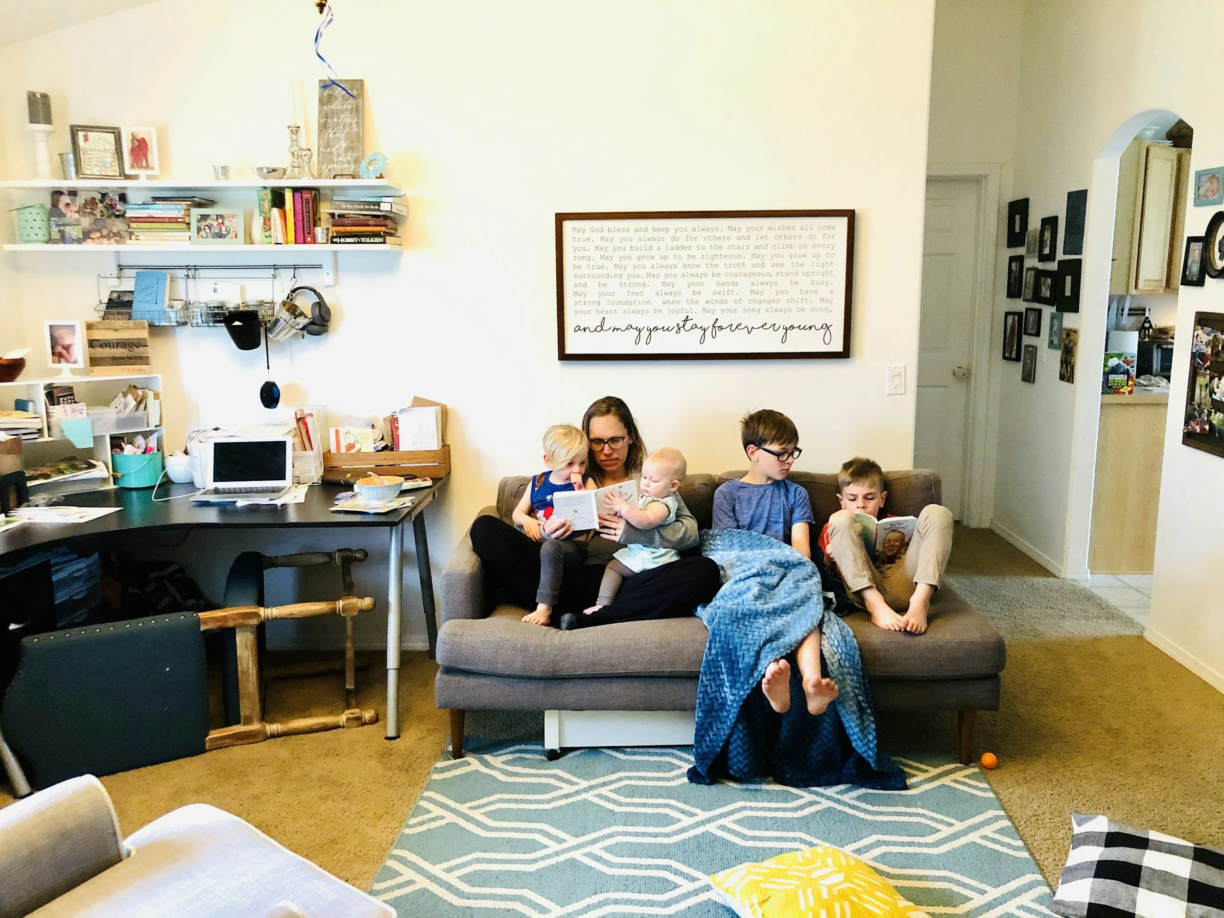 A woman and four children sitting on a gray couch, reading books together in a cozy living room.