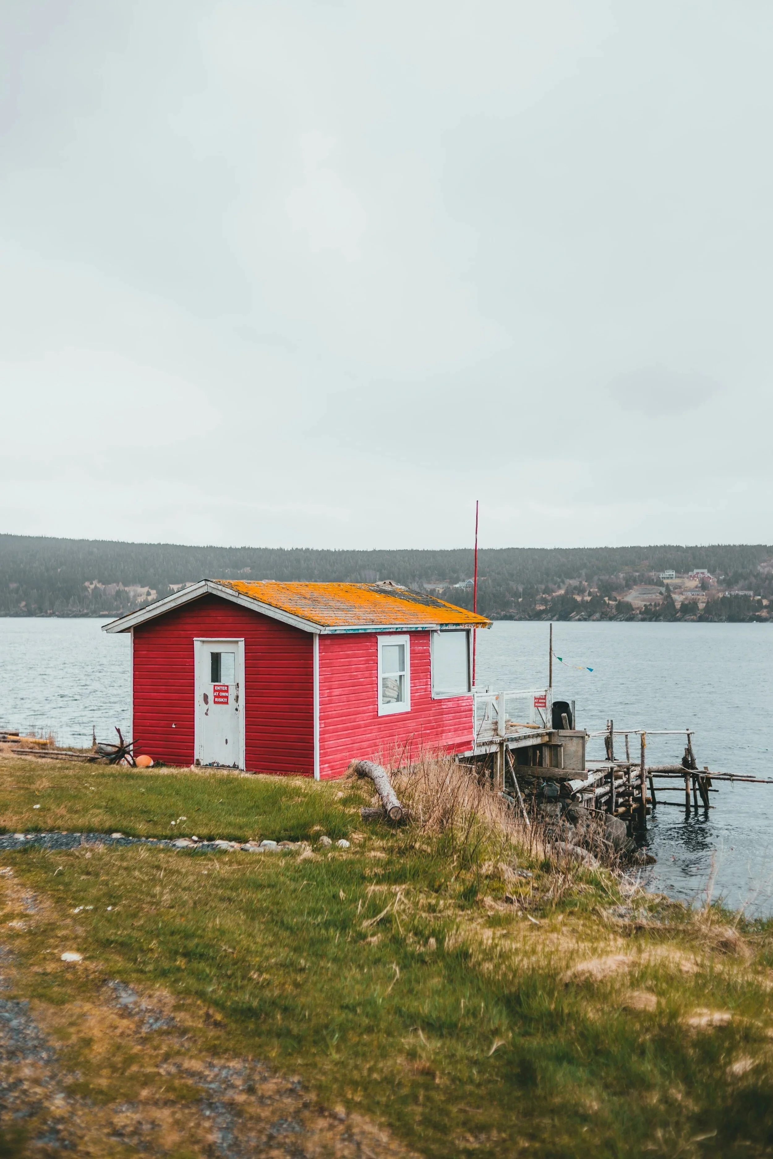A red wooden twine shed on the side of a grassy coastline, leading to a small wooden wharf. The ocean is gray and evergreen hills are visible across the bay.