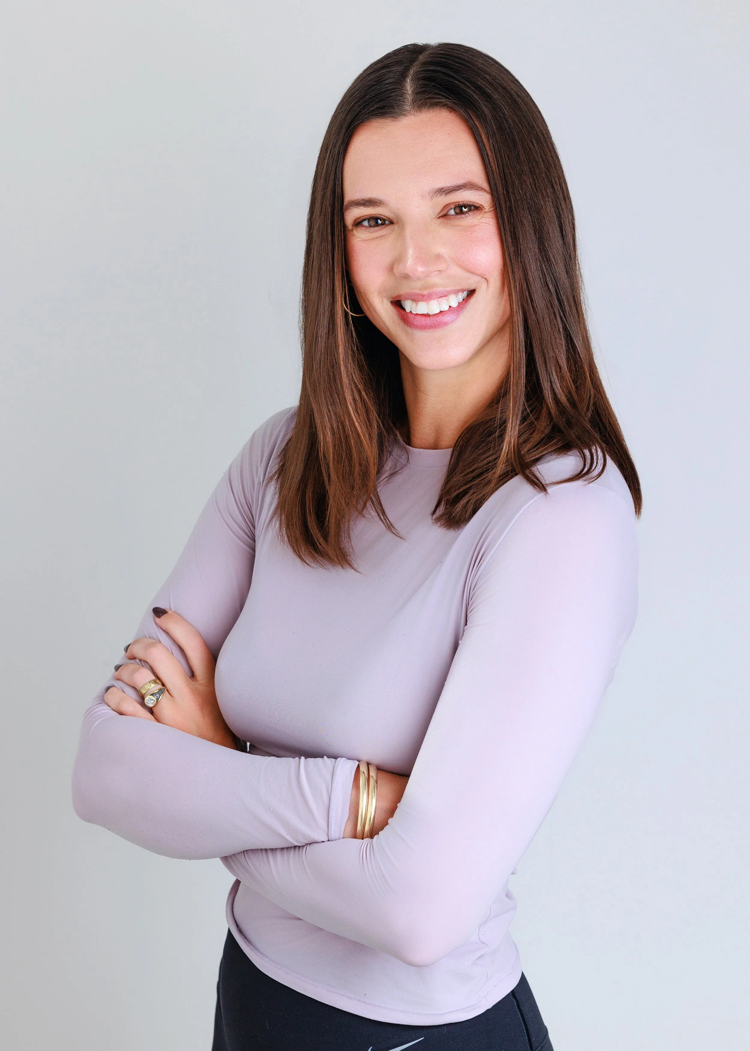 A smiling woman with brown hair and light skin, wearing a long-sleeve lavender top and gold jewelry, standing with arms crossed against a plain light background.