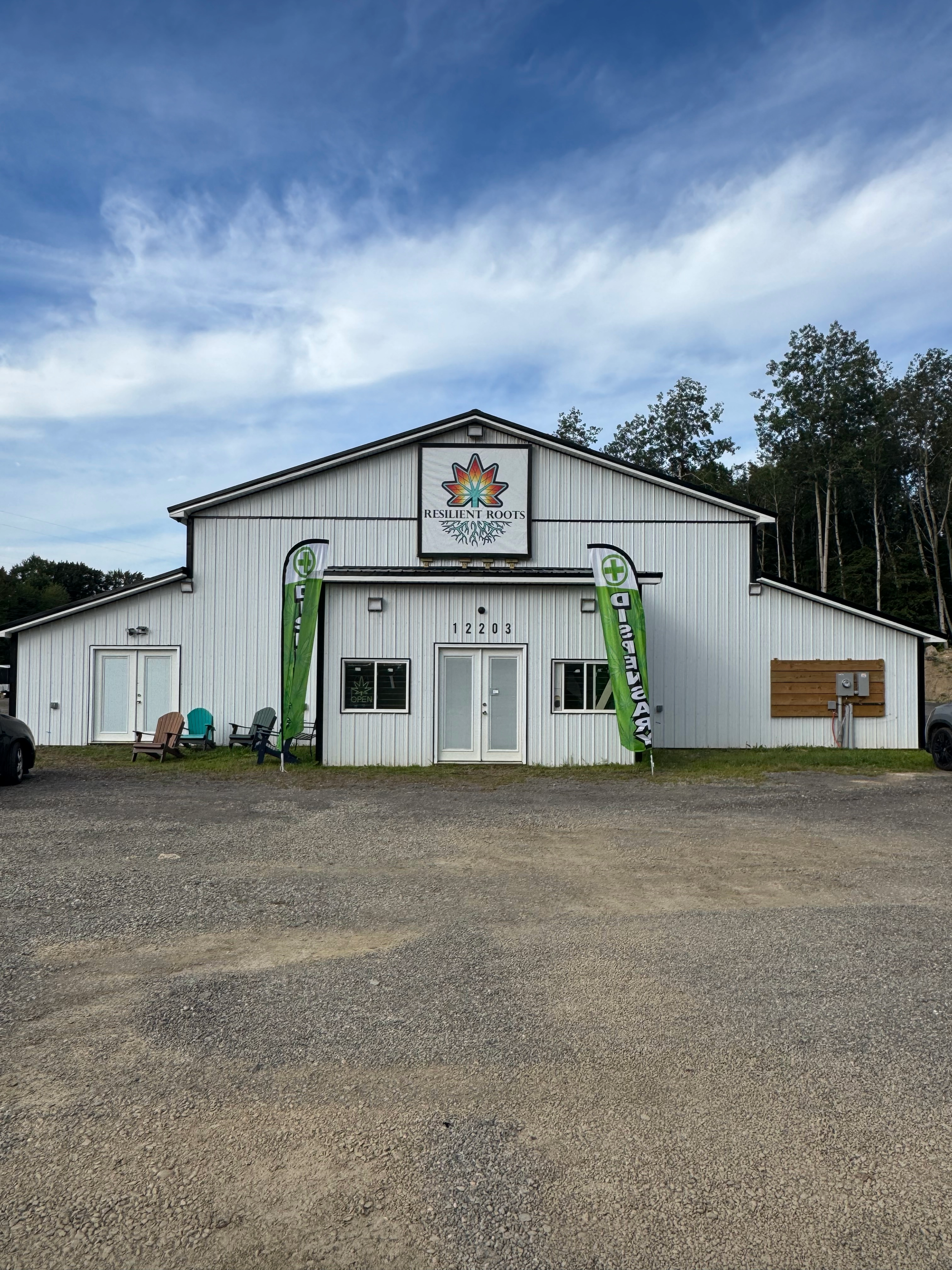 A white building with a black roof, decorated with a colorful maple leaf logo and the words 'Resilient Roots'. There are green flags with white crosses and black text, plastic chairs outside, and a gravel parking area in front. Some trees are visible on a hill behind the building.