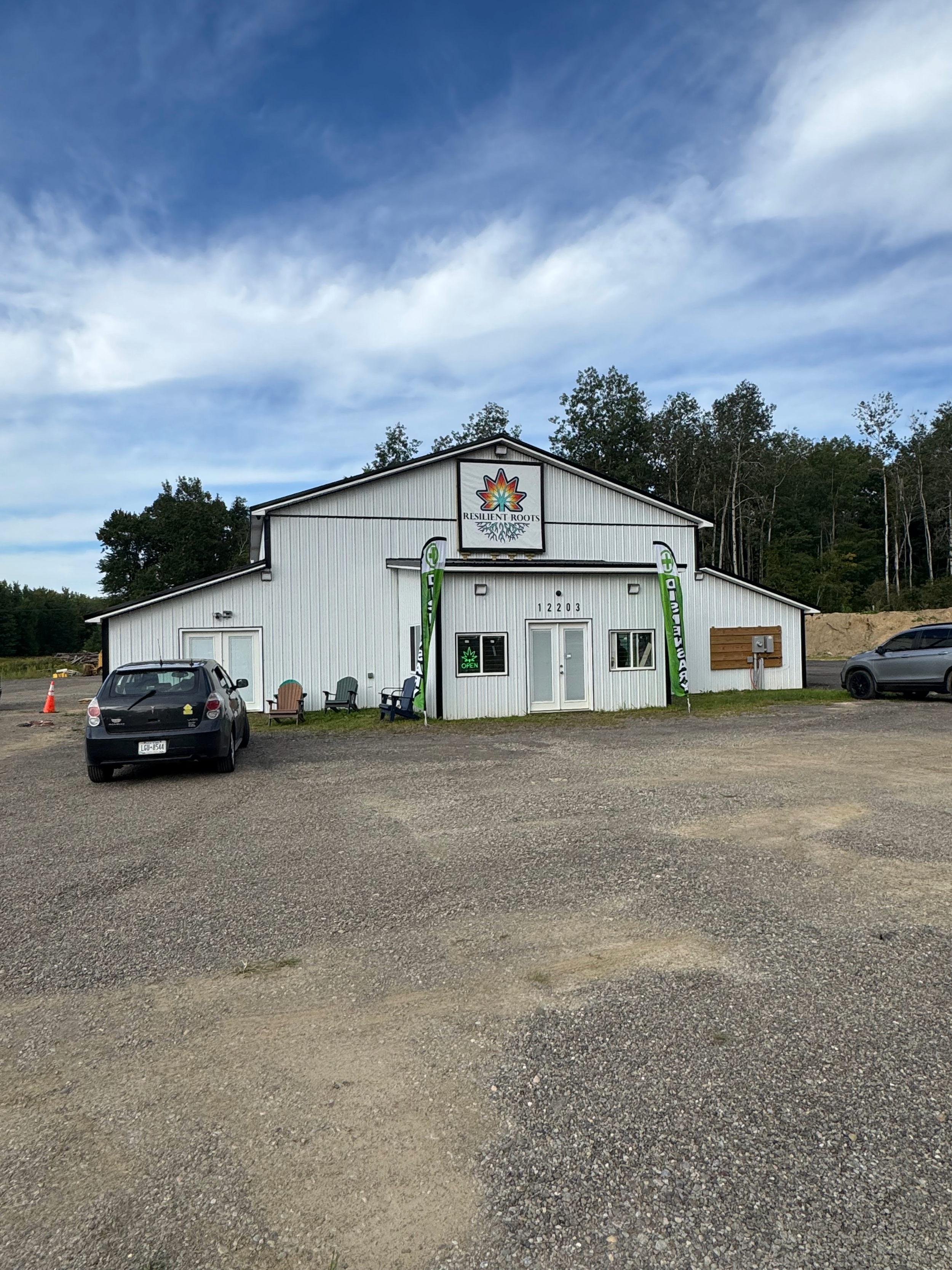 A white building with a sign that reads 'Resilient Roots' and an abstract root and leaf logo, two green vertical flags, two chairs outside, and a parked car, with a background of trees and an open sky.