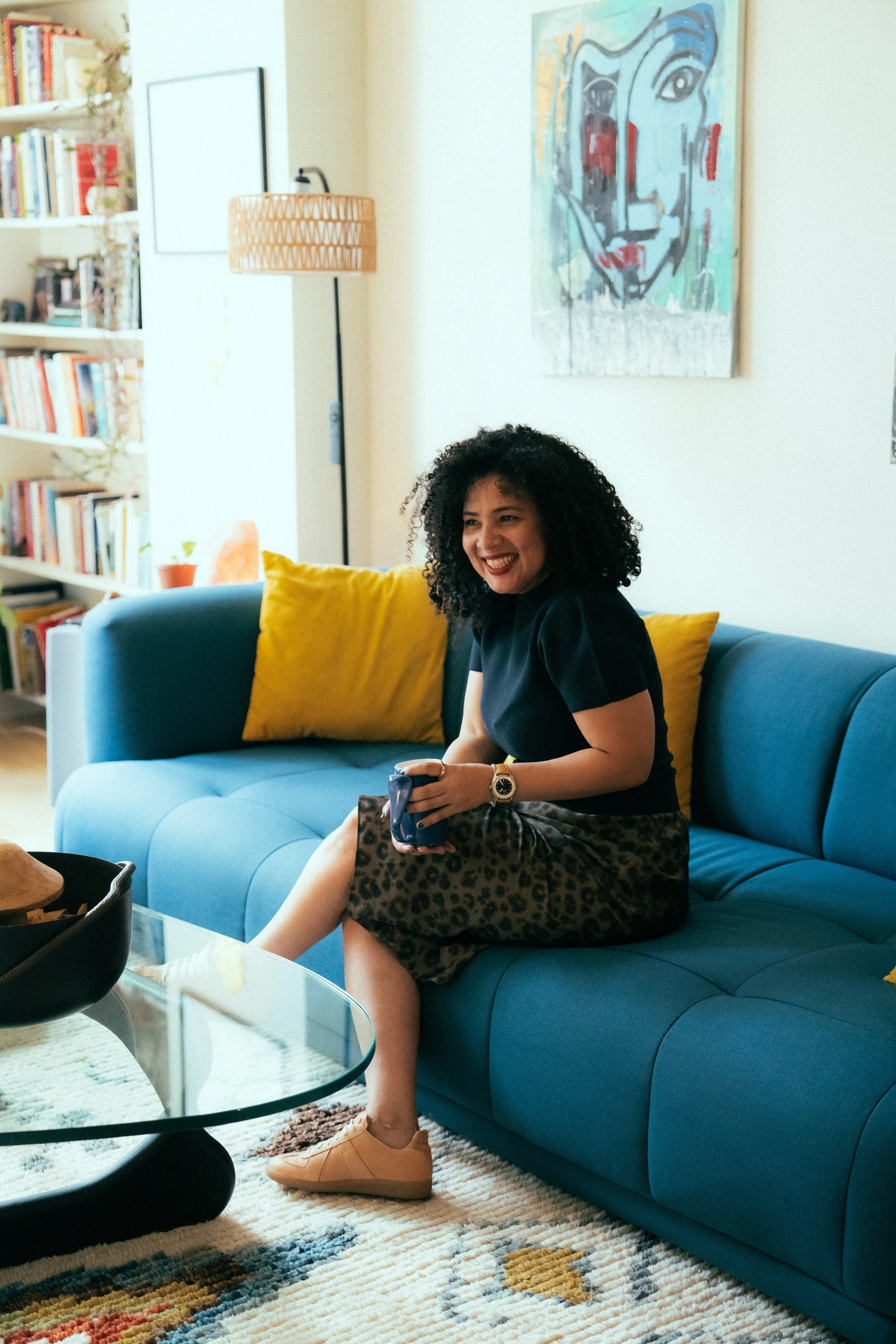 A woman with curly black hair, wearing a black top and leopard print skirt, sitting on a blue couch with yellow pillows. She is smiling and holding a phone. The room has a bookshelf on the left, a painting on the wall, a floor lamp, and a glass coffee table in front of her. There is a multicolored patterned rug on the floor.