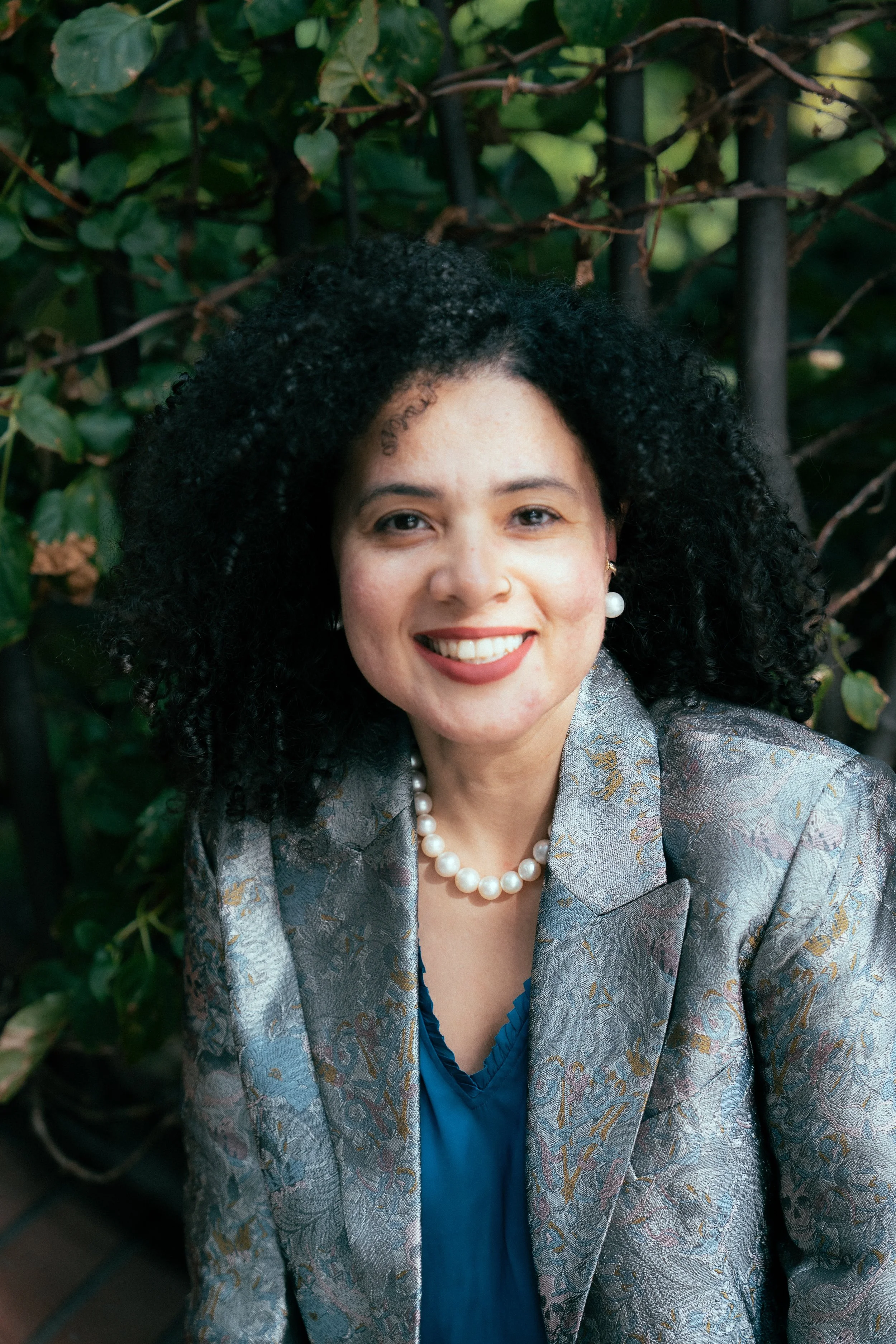 A woman with dark, curly hair smiling, wearing pearl earrings, a jade-colored ruched dress, and a silver patterned blazer, standing outdoors with green foliage in the background.