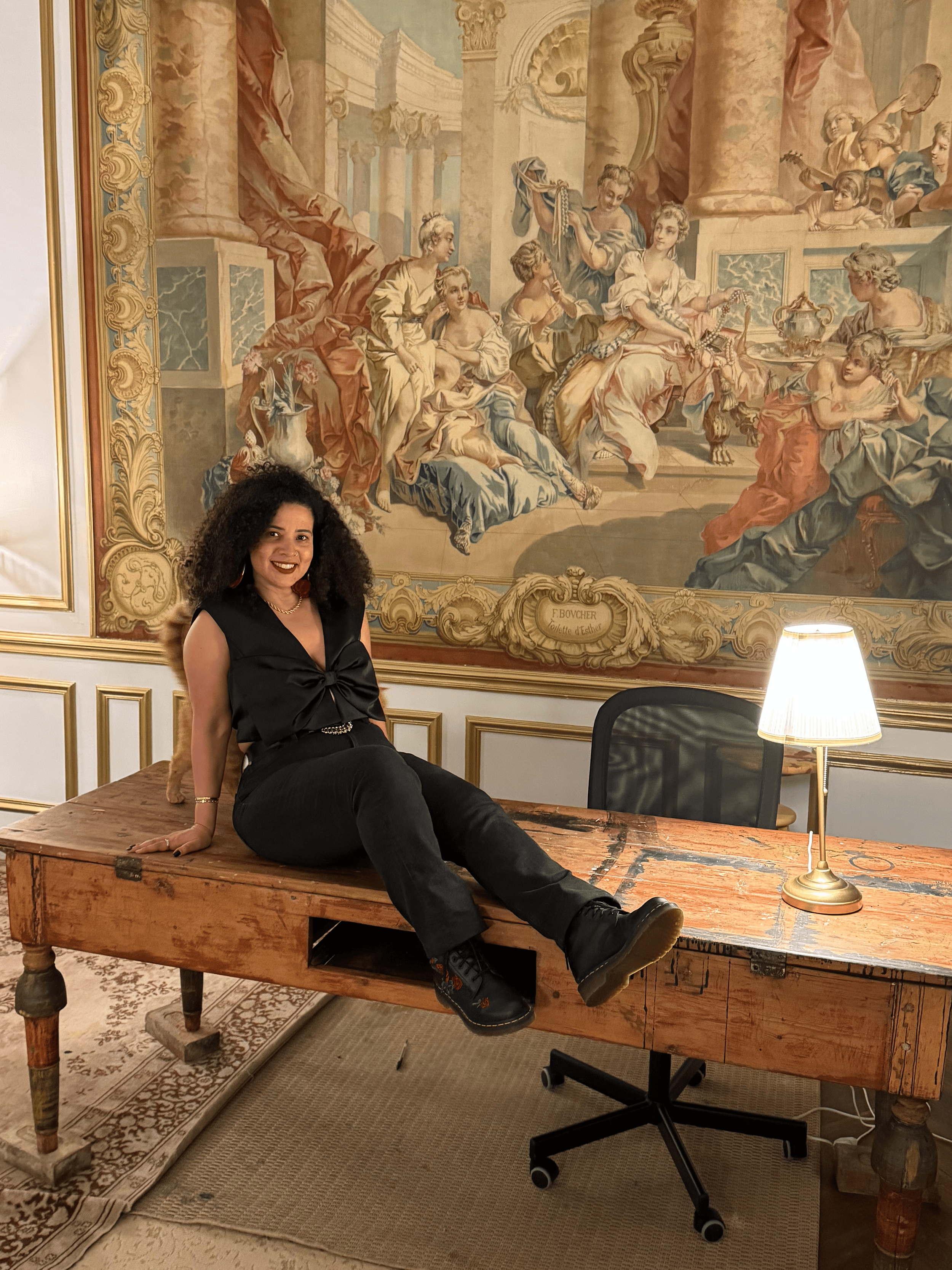 A woman with dark curly hair wearing a black sleeveless top and black pants sits on a vintage wooden table with one leg extended and the other bent. Behind her is a large classical style painting depicting a scene with multiple women and children in a palace setting, with pillars and ornate decorations. To the right, there is a small table lamp with a beige shade illuminating the scene. A rolling office chair is partially visible in front of the table.