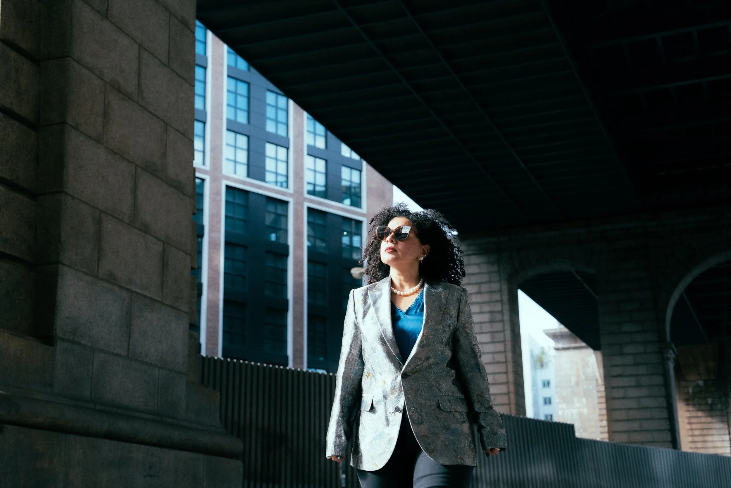 A woman with curly dark hair wearing sunglasses, a pearl necklace, a patterned blazer, and a blue top walking under a bridge in an urban area with tall buildings.
