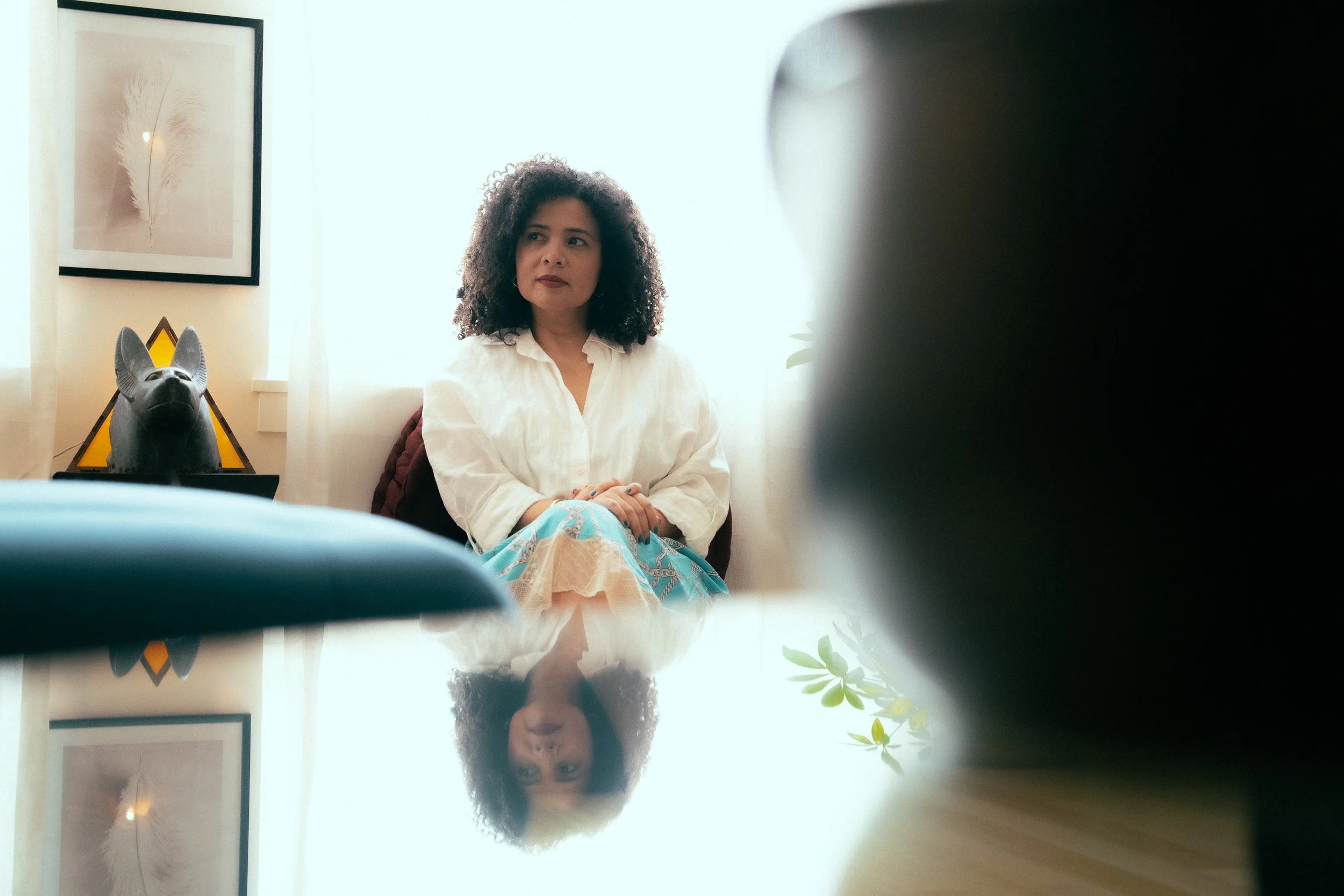 A woman sitting in a well-lit room with framed artwork and a decorative dog head sculpture on a table, as seen through a mirror or reflective surface.