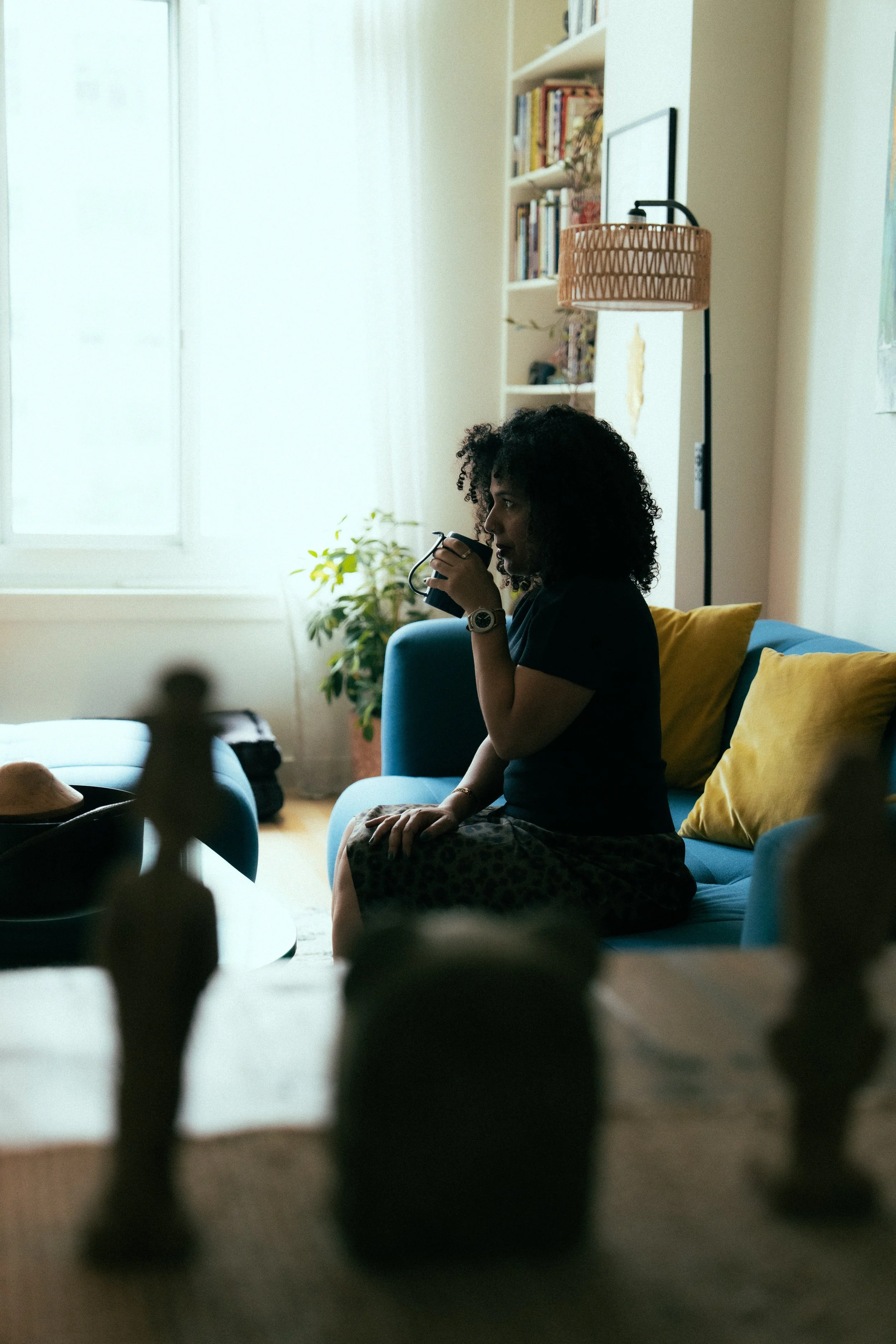 A woman with curly hair sitting on a blue couch, holding a mug in her right hand, in a cozy living room with a bookshelf, plants, and a floor lamp.
