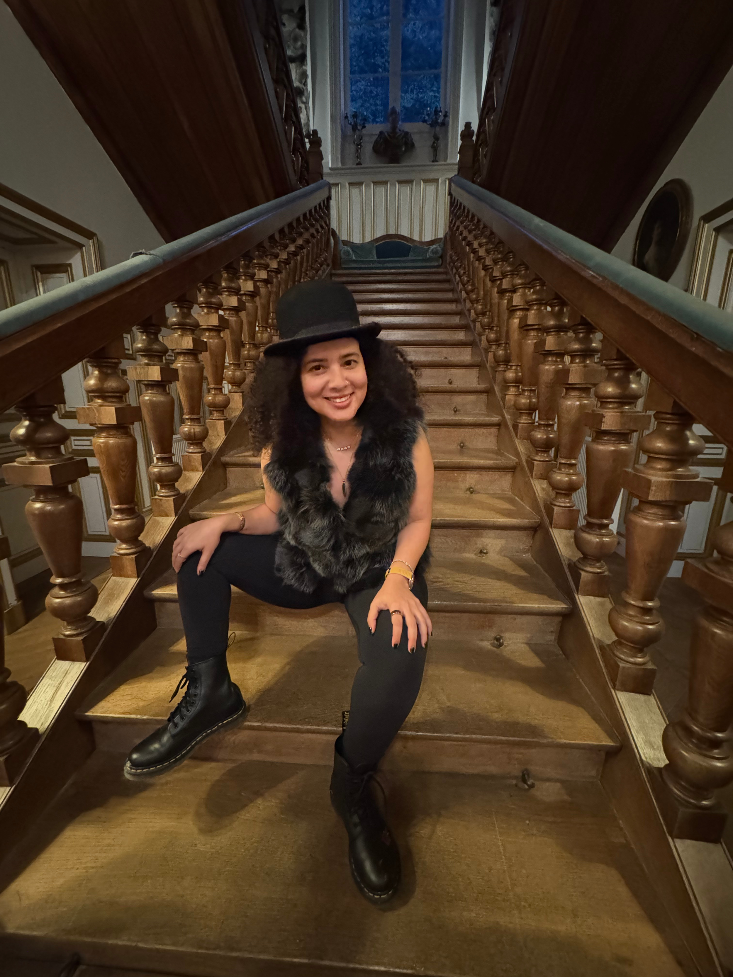 A woman with curly hair dressed in black with a fur vest, wearing black combat boots, sitting on wooden stairs in a historic building, smiling at the camera, with a window and decorative elements in the background.