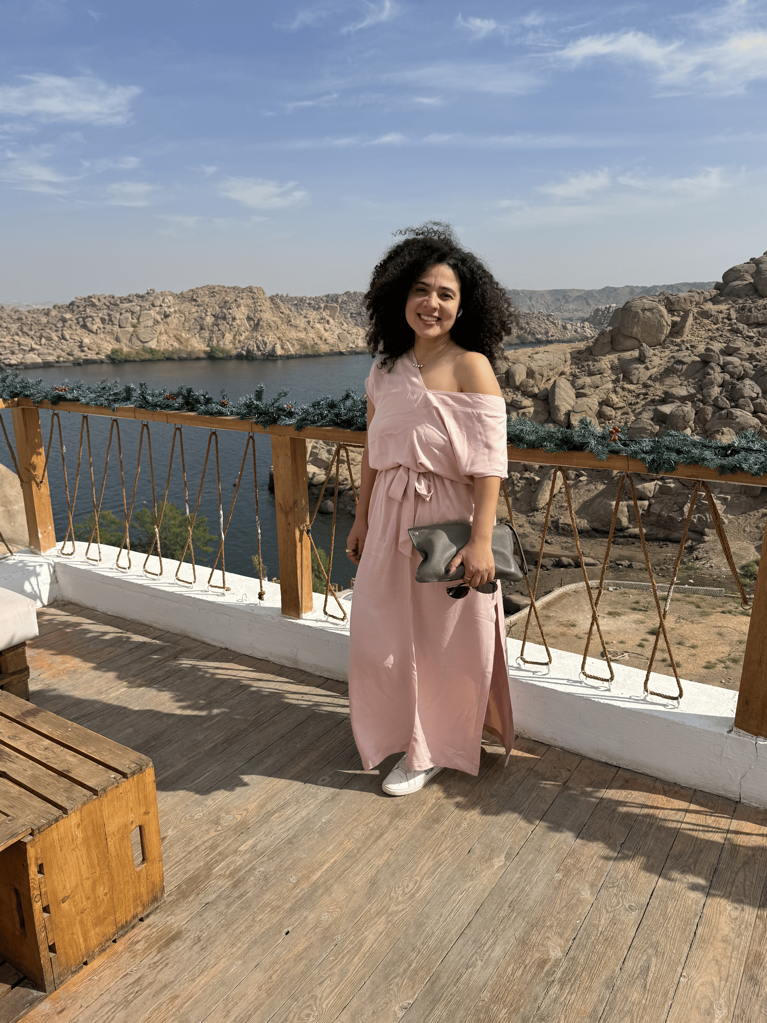 A young woman in a pink dress holding a gray clutch, standing on a wooden deck with a scenic view of a body of water and rocky hills in the background, smiling at the camera.
