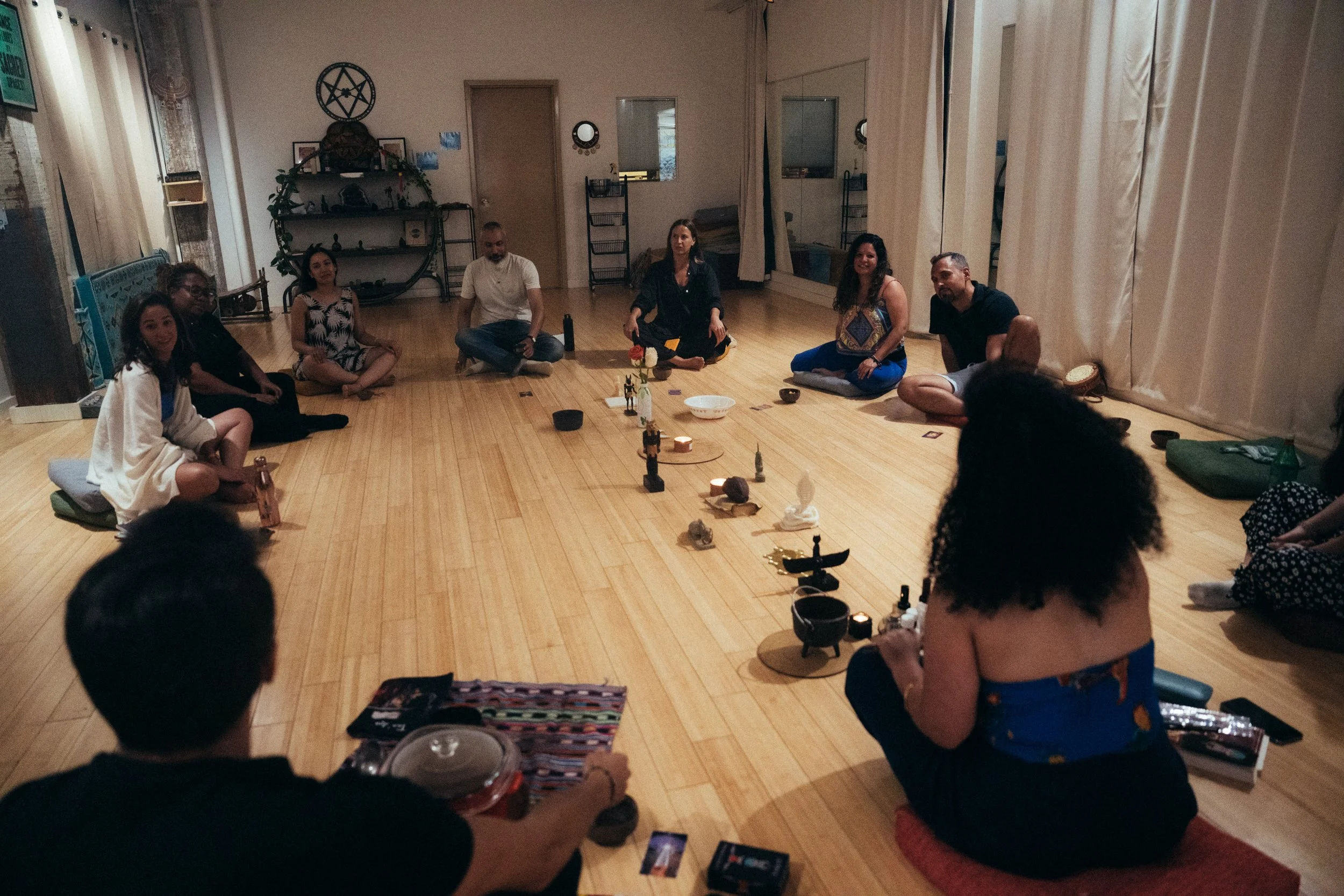 A group of people sitting in a circle on the floor in a room with wooden floors, engaged in a meditation session with various objects and candles on the ground in front of them.