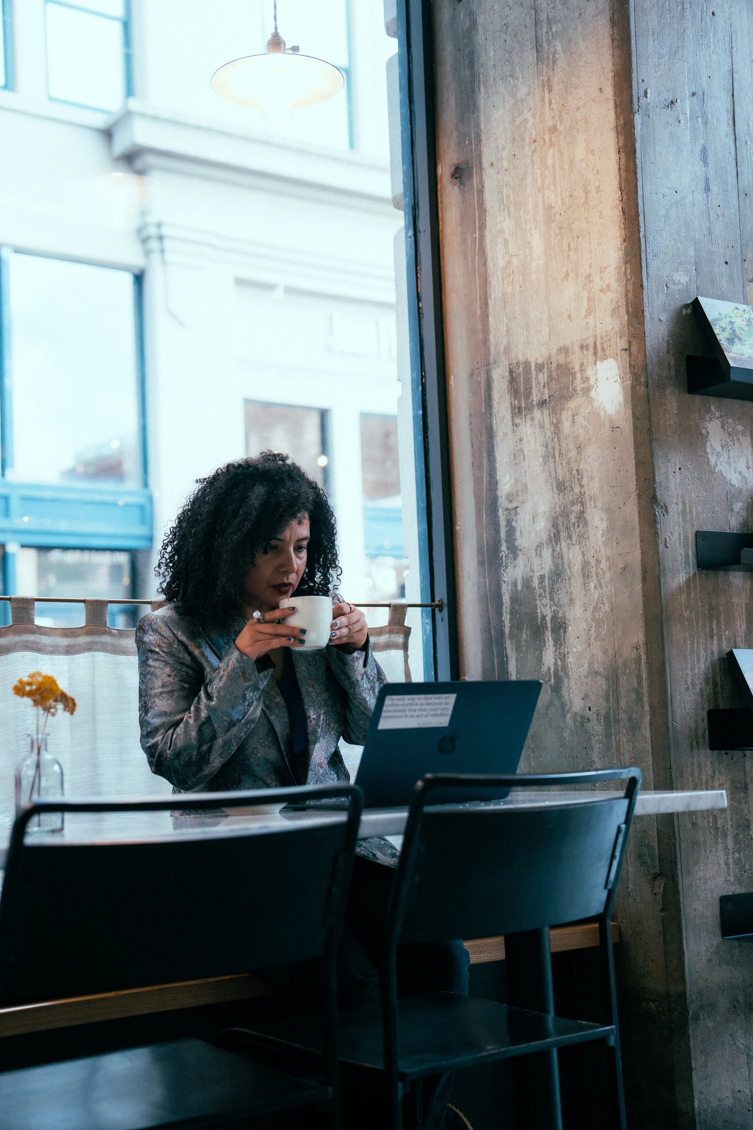 A woman with curly black hair sitting at a table in a café, holding a white mug, with an open laptop in front of her and a flower in a vase on the same table. The café has a rustic exposed brick wall and large windows letting in natural light.