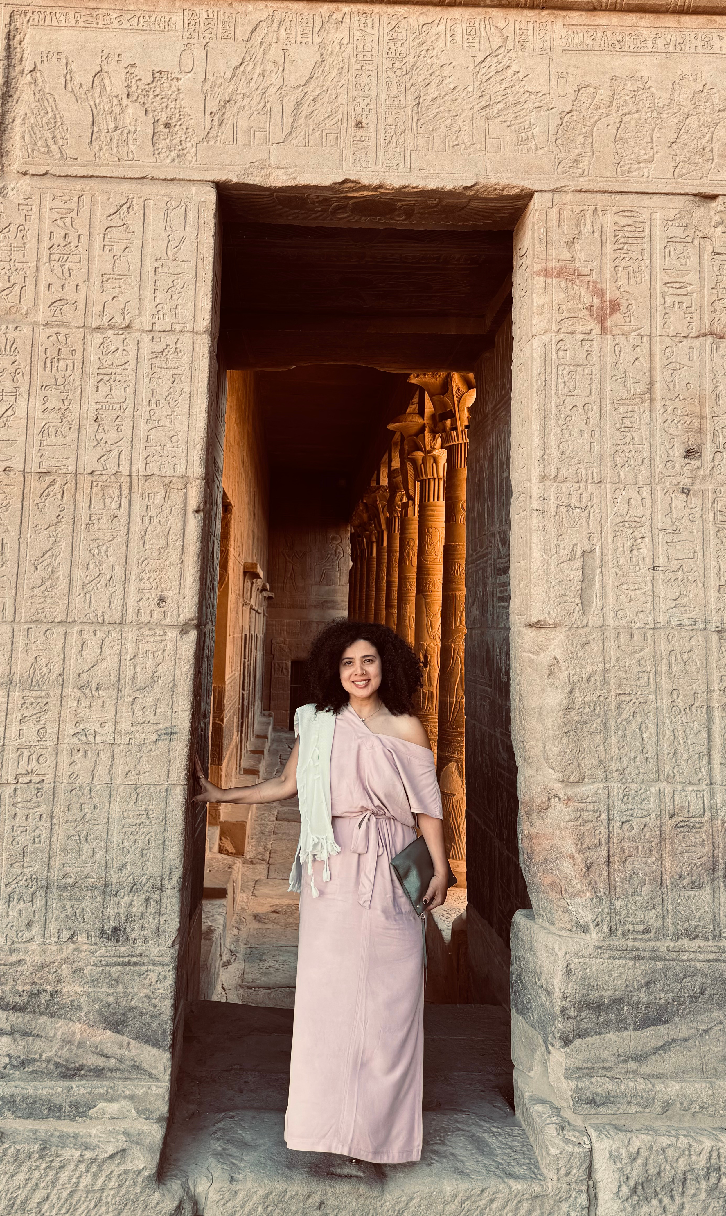 A woman in a pink dress and light-colored shawl standing in the doorway of an ancient Egyptian temple with hieroglyphics on the stone walls and yellow columns with carvings inside.
