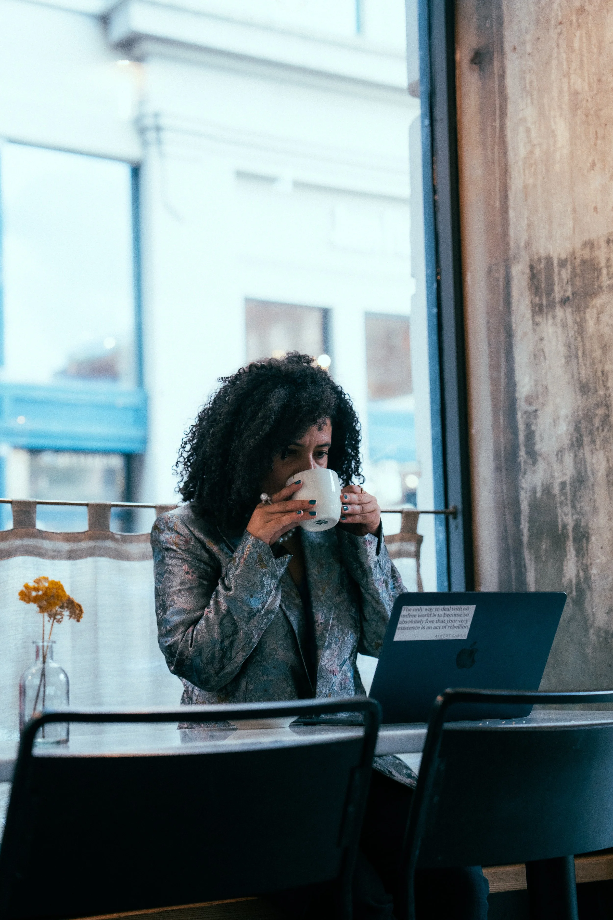Woman with curly hair wearing a patterned blazer, sitting in a cafe, drinking from a white mug while working on a laptop.