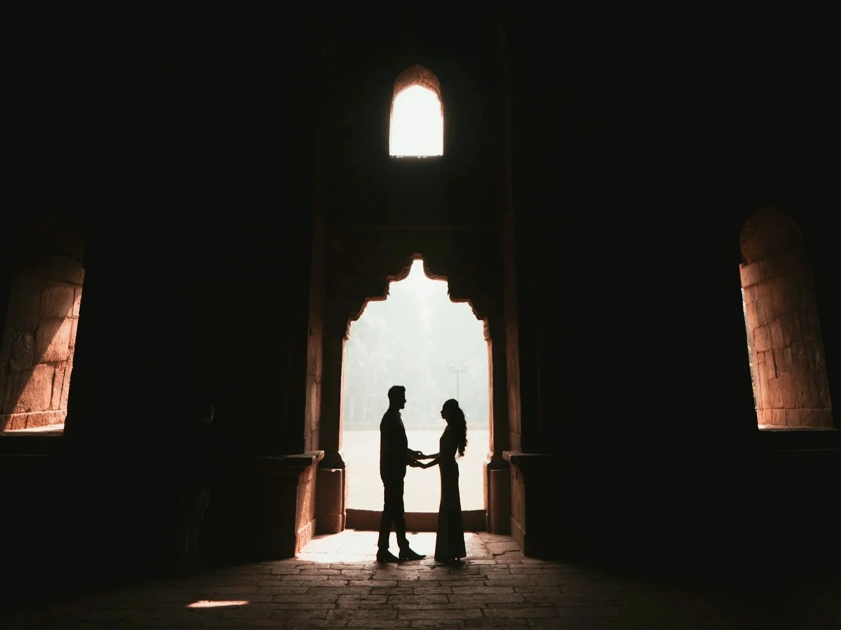 Silhouette of a couple holding hands inside an arched doorway of a historic building, with natural light illuminating the scene from outside.